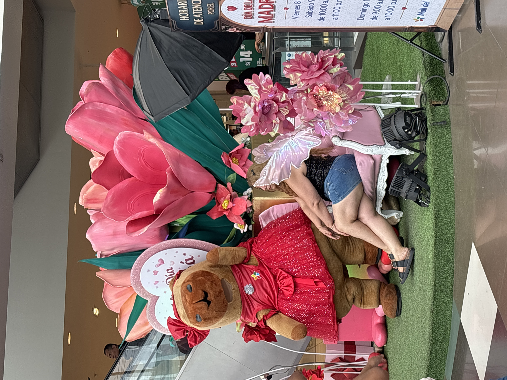 The image shows a Mother's Day display at a mall in San Juan de Miraflores, Peru. The display features a large floral arrangement with pink flowers and green leaves, a teddy bear wearing a red dress, and a sign with information about Mother's Day events. A woman in a denim skirt and black shirt is seated on a white chair next to the teddy bear, posing for a photo. The floor is covered in artificial grass. Electrical lights can be seen around the chair. A sign overhead advertises hours and special deals at the mall. A partial view of a pedestrian escalator is seen to the left of the display.