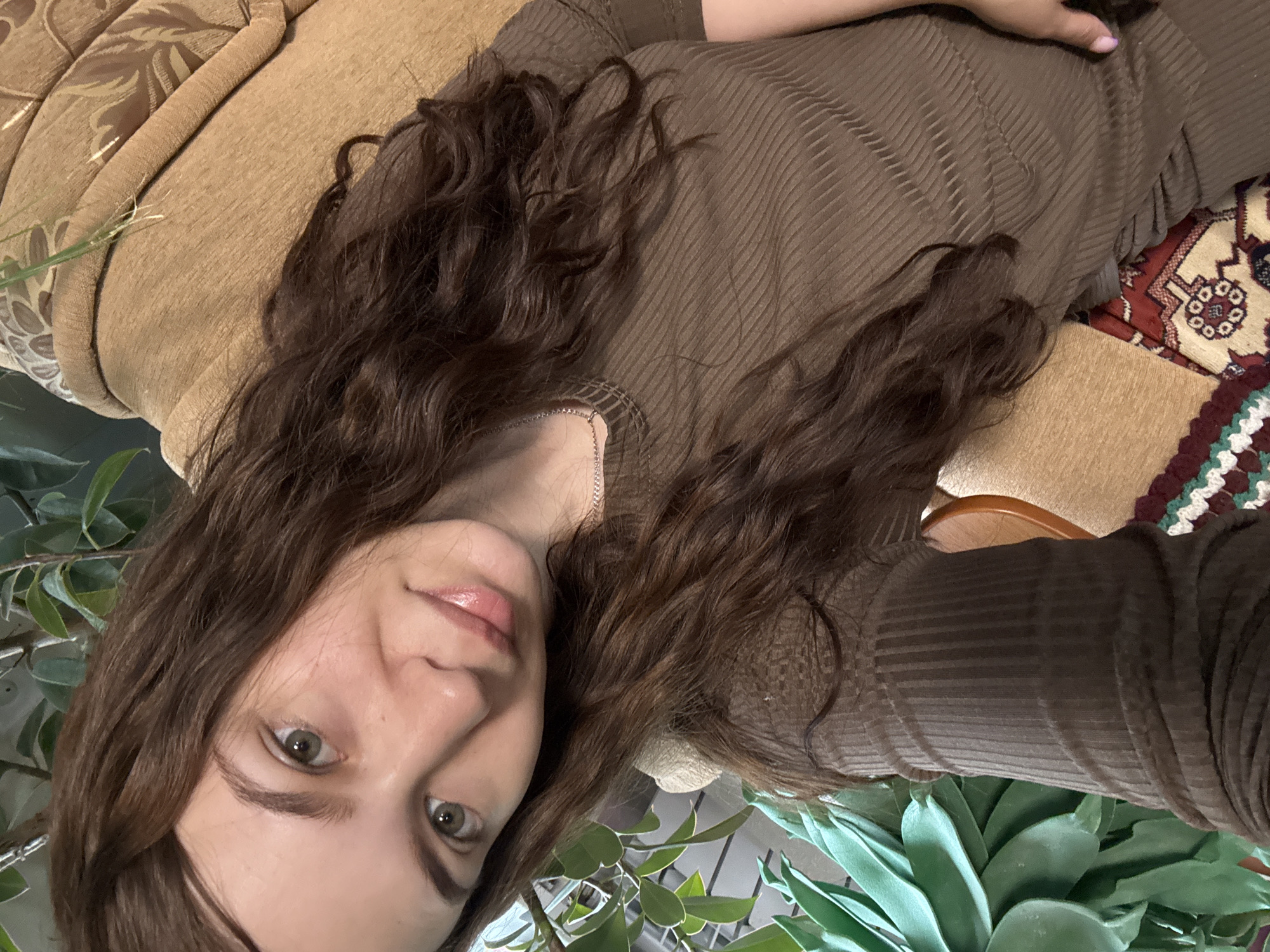 A woman with brown curly hair, fair skin, and green eyes is shown in a selfie-style shot. She is wearing a brown ribbed top. Her hair is partially draped across the fabric. She is positioned on or near a beige patterned couch. Plant life, with green leaves, is visible in the foreground. A patterned rug is partially visible in the background. The location information is irrelevant to the visual content of the image.