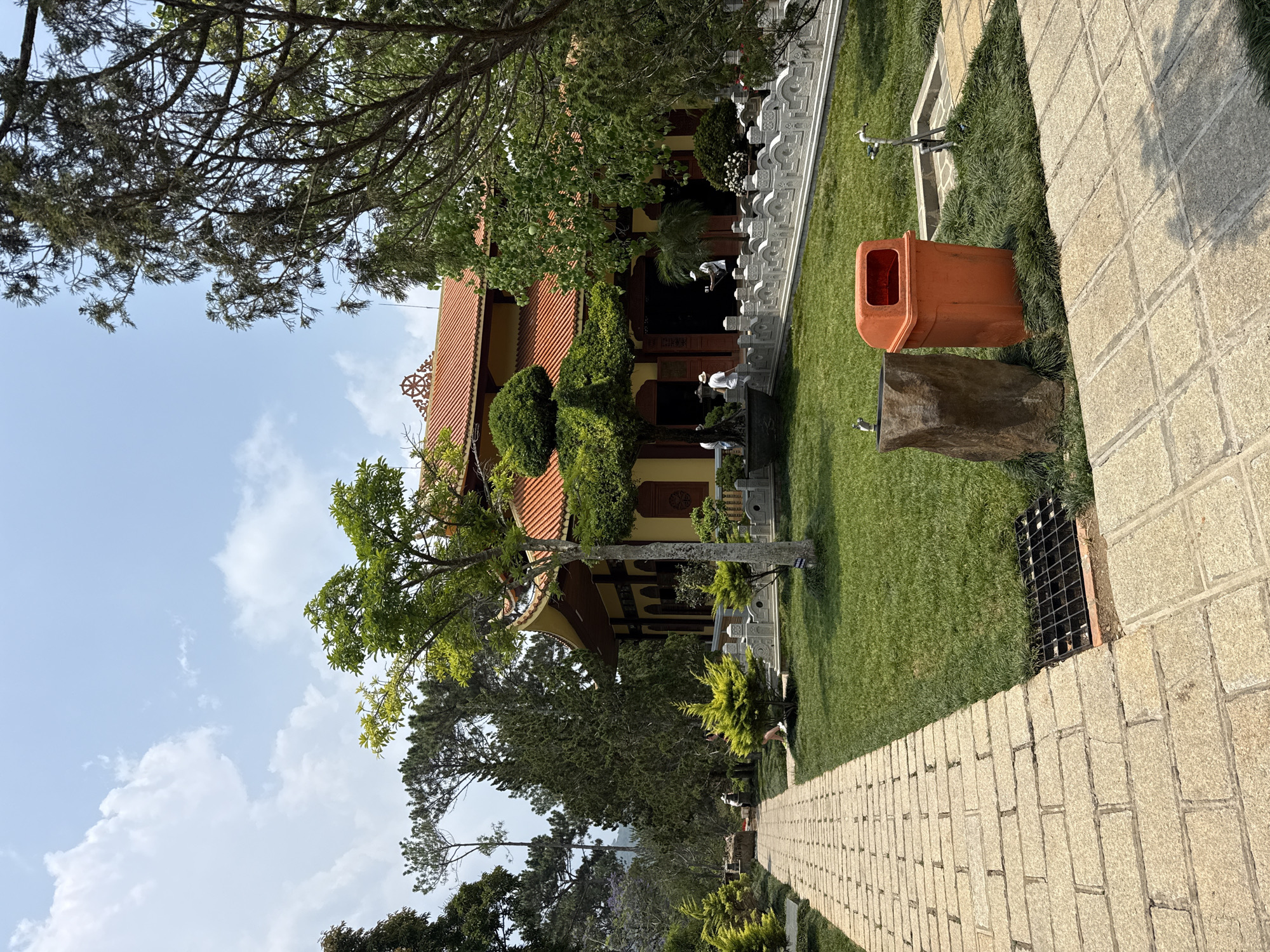 The photo shows a temple in Da Lat, Vietnam, viewed from an eye-level perspective along a stone-paved path. The temple is a two-story structure with a terracotta-colored tiled roof, a facade adorned with detailed carvings, and landscaped with shrubs and potted plants. A decorative white stone railing runs along the edge of the temple's courtyard. An orange trash receptacle stands on a manicured lawn near the path, positioned next to a rock-like water fountain. The path stretches into the distance, flanked by grass, trees, and low-lying bushes. The sky is light blue, dotted with scattered white clouds. The overall impression is one of serenity and well-maintained grounds.