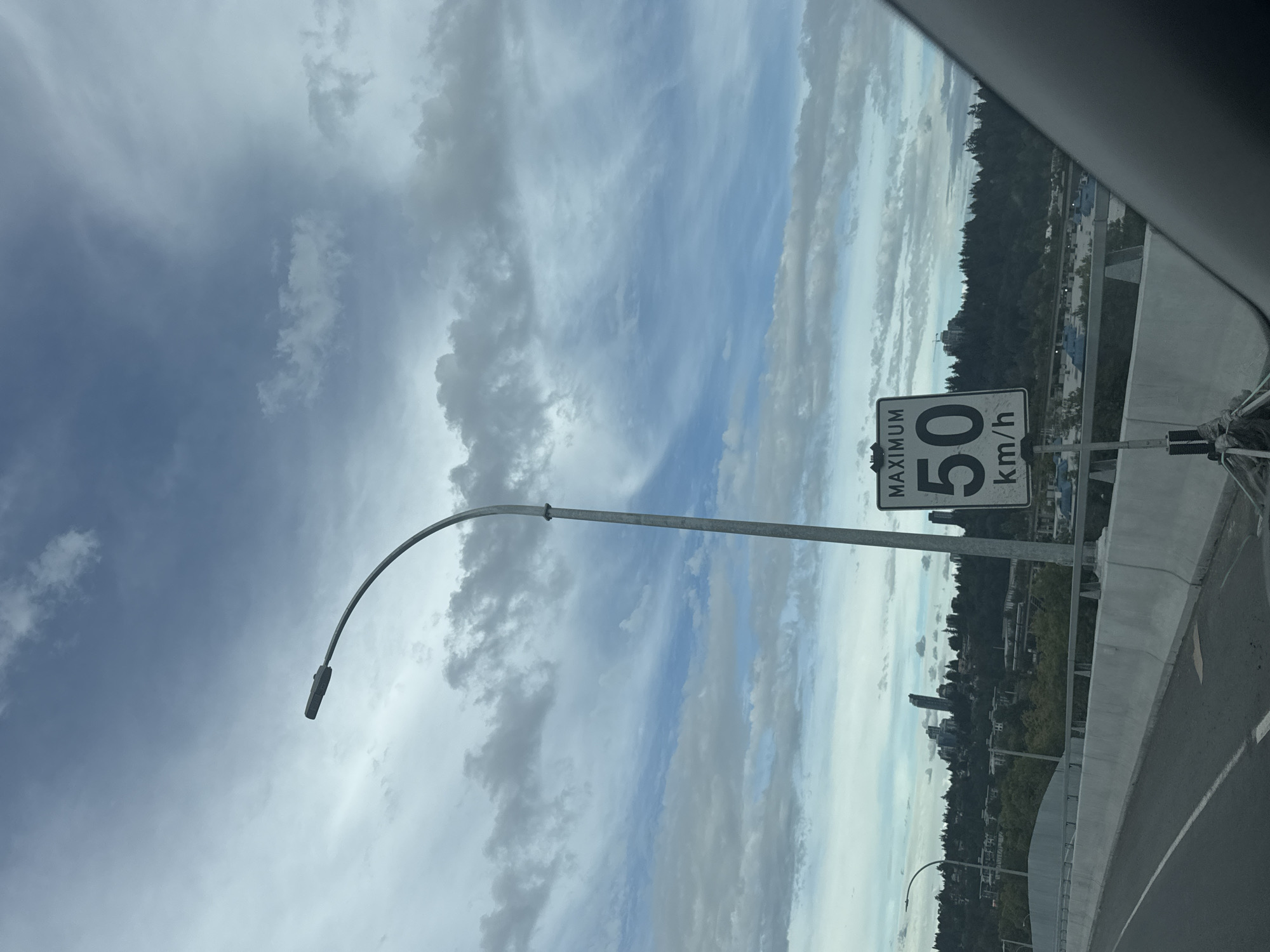 This is a photograph taken from a vehicle on a highway overpass, likely the Pattullo Bridge, in Surrey, Canada. The sky is partly cloudy with dramatic, elongated cloud formations. In the foreground, a concrete barrier with a white stripe runs along the right side of the road. A street light with a curved arm extends over the road. A black and white speed limit sign is prominently displayed, indicating "MAXIMUM 50 km/h." Beyond the overpass, a dense line of trees forms a natural border against the horizon, with buildings and a few taller structures, suggesting a cityscape, visible in the distance. The overall lighting suggests an overcast or late afternoon sky. There are no people visible in the image.