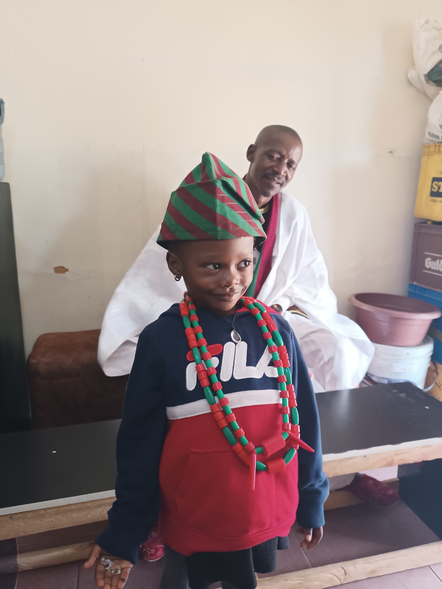 An indoor portrait captures a young child and an adult man in Jos, Nigeria. In the foreground, a dark-skinned child stands with a gentle smile, looking slightly off-camera to the right. The child wears a distinctive green and red striped conical hat, multiple strands of red and green beaded necklaces, a dark blue and red hoodie with "FILA" emblazoned in white on the chest, and black leggings. A small hoop earring is visible in the child's left ear, and their left hand is open, holding small, light-colored fragments.
Behind and slightly to the left of the child, an adult man is seated on a brown sofa, observing the child with a warm, benevolent smile. He is dark-skinned, balding, and dressed in a traditional white robe with a red/maroon trim around the neck. The background features light-colored walls and a functional environment, including a yellow crate with "STAR" partially visible, other containers like white buckets and a reddish-brown plastic basin, and a white bag hanging on the wall. The floor is covered with light-colored tiles. The bright, even lighting suggests a daytime setting. The scene conveys a sense of cultural celebration or a special occasion, marked by affectionate interaction between the subjects.