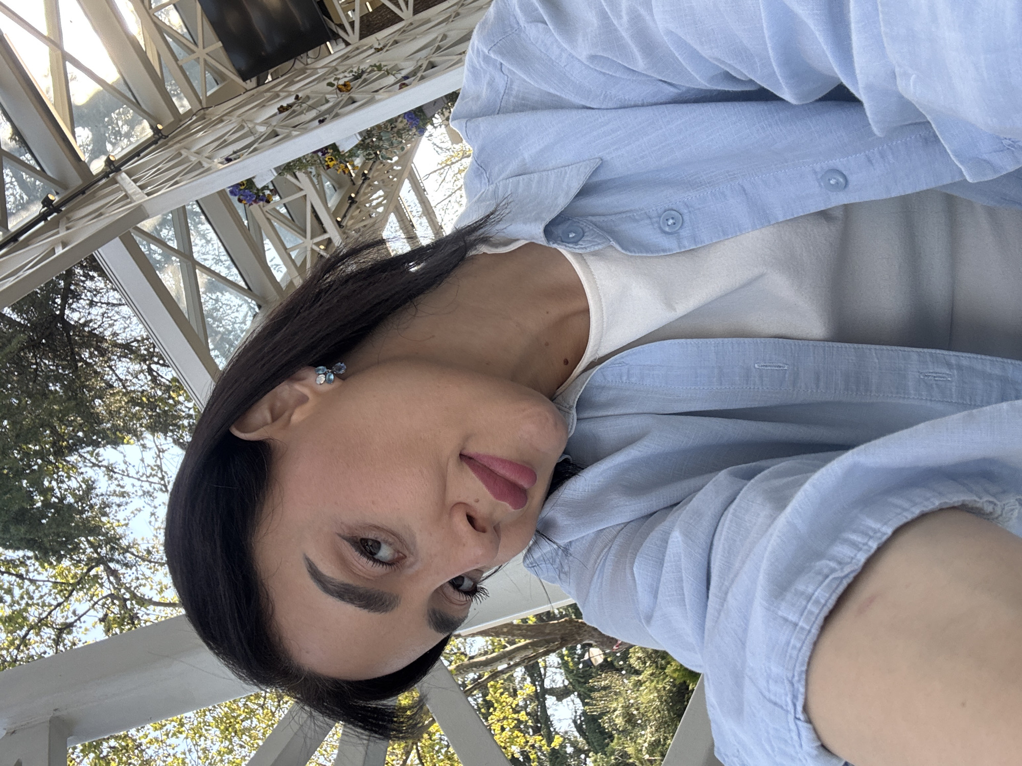 The image features a woman posing for a close-up, selfie-style photograph. She has dark hair, styled to fall over her shoulders, and pronounced dark eyebrows. Her eyes are looking directly towards the camera, and her lips, painted with dark pink or mauve lipstick, are slightly parted, conveying a calm or subtly content expression. She wears a light blue, long-sleeved button-down shirt, unbuttoned over a white top. On her visible ear, she sports a small, intricate earring with light blue and silver elements, possibly floral in design.
The background depicts an outdoor setting with a decorative white architectural structure overhead, possibly a pergola or veranda, characterized by crisscrossing beams and supports. Small potted plants or flowers with purple and yellow blooms are visible integrated into this structure. Beyond, lush green foliage and trees are prominent, indicating a natural, garden-like environment, potentially a park or an outdoor cafe. The scene is brightly lit by natural daylight. There is no visible text in the image. Given the provided context, the location is Tbilisi, Georgia.