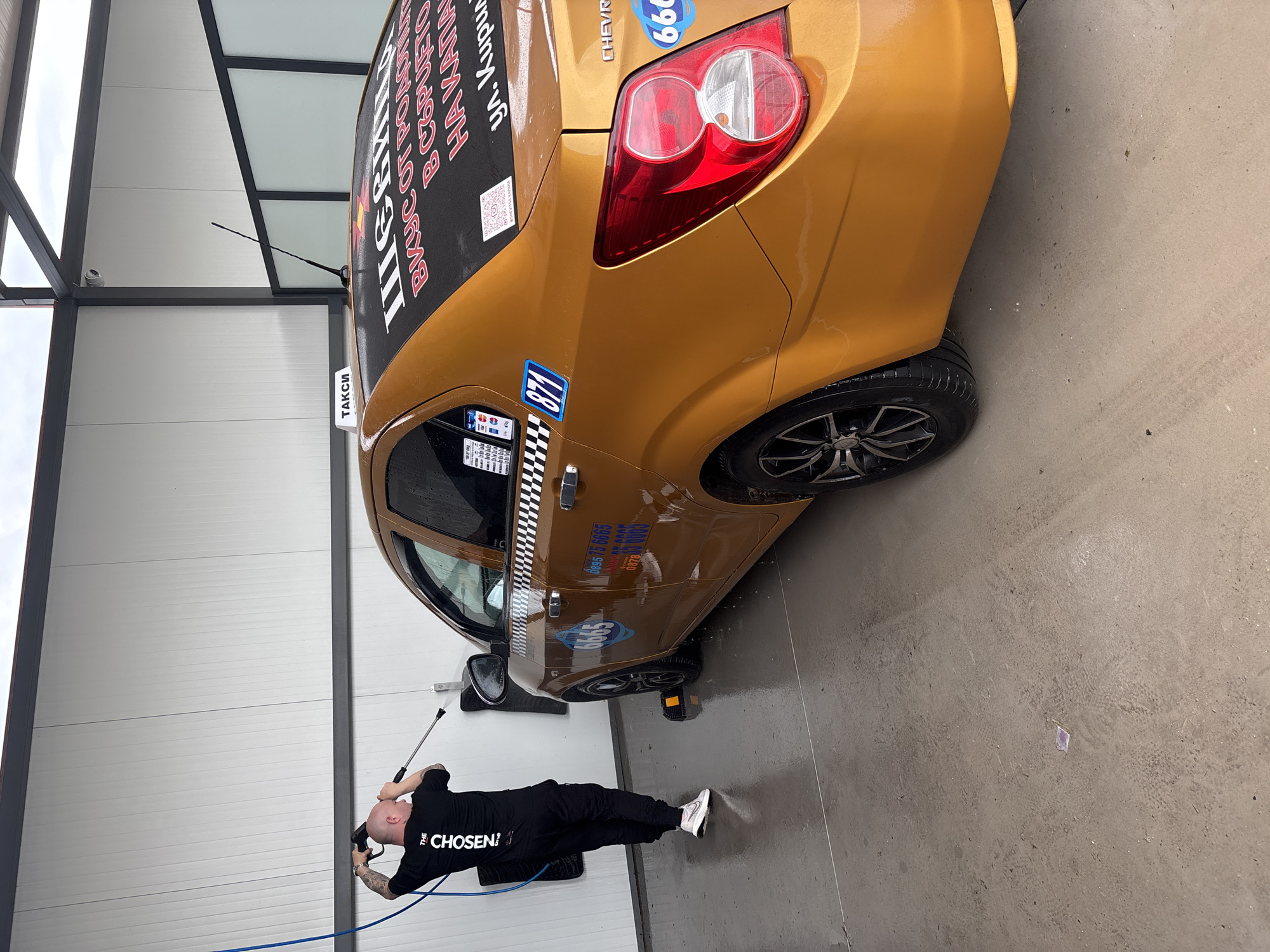 The image shows a man power washing a golden-colored taxi in what appears to be a car wash bay. The man, dressed in black with the word "CHOSEN" printed on his shirt, is holding a pressure washer wand, directing the spray towards the vehicle. The taxi has advertising decals and stickers on it, including a checkered pattern and a "TAKCИ" (Taxi) sign. The background is a wall made of corrugated metal panels, and the floor is wet concrete. The information on the taxi suggests that the car is located in Novo Selo, Bulgaria.