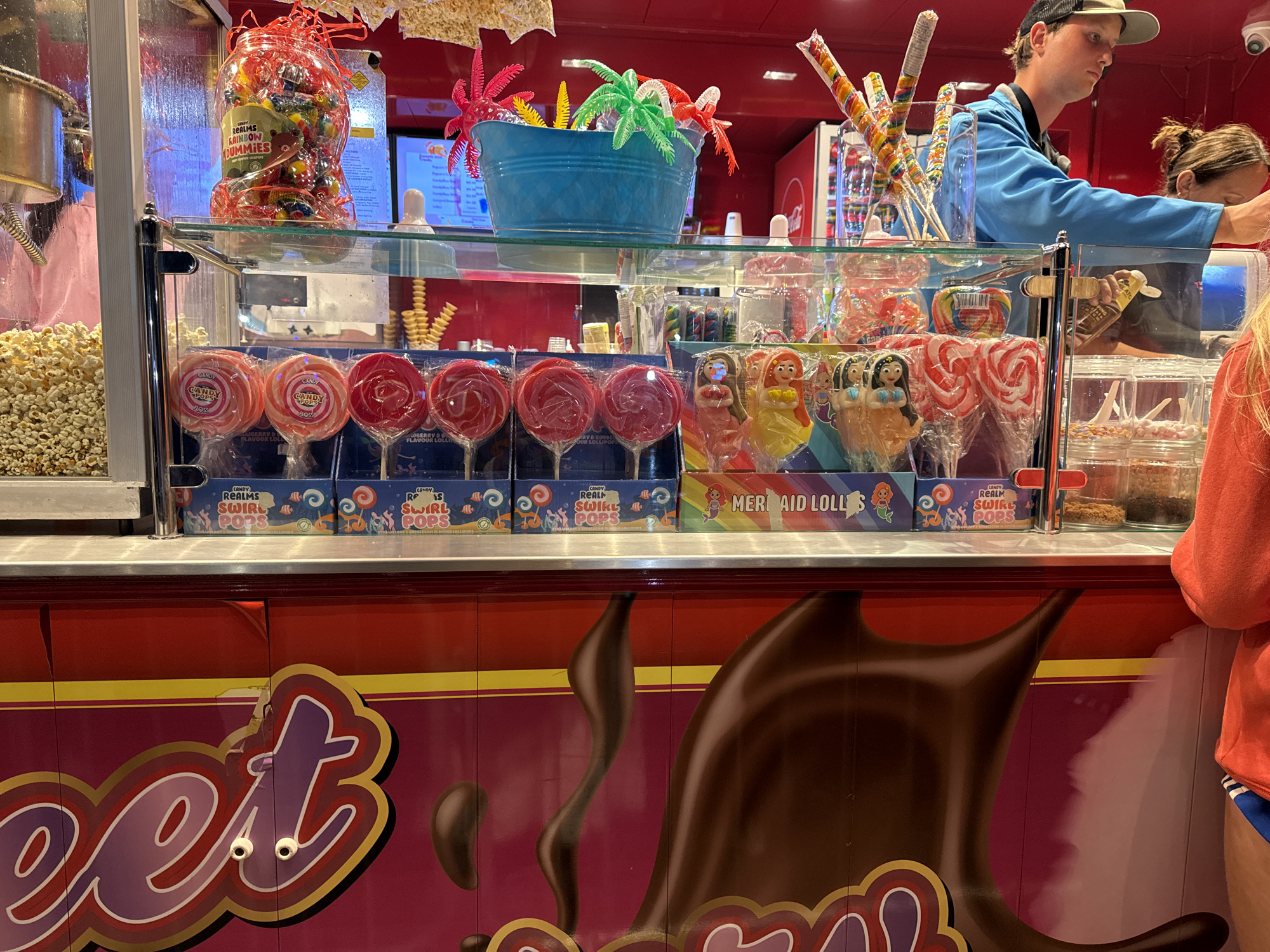 This image captures a close-up view of a candy stall, likely situated in Tramore, Ireland, given the context provided. The stall features a vibrant display of sweets, with a primary focus on lollipops and other sugary confections.
Dominating the foreground is a large, red-colored candy cart with a design that evokes melted chocolate and dripping icing, further enhanced by the prominent word "Sweet" written in stylized purple lettering with a yellow outline. The cart has two circular white objects resembling googly eyes, adding a whimsical touch to the overall aesthetic.
Behind the cart, a glass display case showcases an assortment of candies. On the left, a large glass jar filled with colorful, wrapped gummy candies is visible. Below this, a row of pink and red swirl lollipops is neatly arranged in their packaging. Further to the right, a collection of character-shaped lollipops, including mermaids and other figures, are displayed. There are also clear plastic containers filled with various types of small candies.
In the background, a person wearing a blue jacket and a cap is visible, seemingly attending to the stall. Another person, wearing an orange top, is also present. A popcorn machine can be seen on the far left, with a batch of popcorn displayed. The stall is decorated with bright colors and playful elements, typical of a fairground or amusement park setting. The overall impression is one of a cheerful and abundant candy emporium.