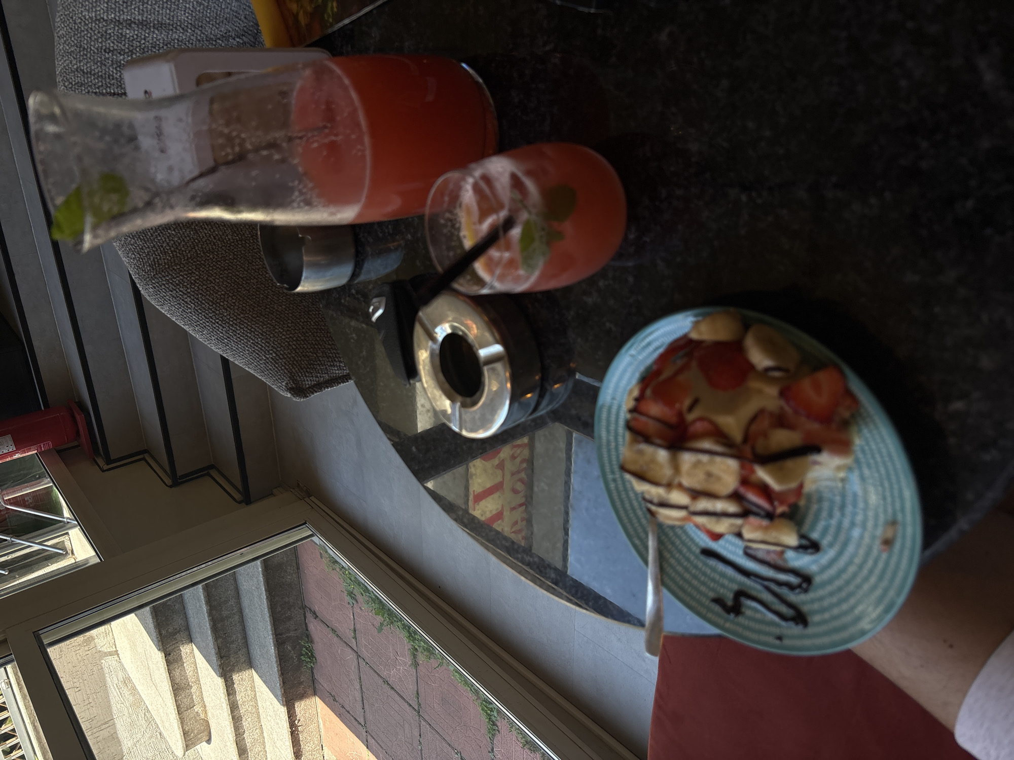 The image depicts a table in what appears to be an outdoor cafe setting, likely in Plovdiv, Bulgaria, given the context of the user's request for a location in that city. The table is made of dark, speckled material, possibly granite or a similar stone, and is situated near a large window that offers a view of an outdoor staircase and tiled patio.
On the table, there are several items:
- A light blue plate with a textured pattern, holding a waffle or pancake topped with sliced strawberries and bananas, drizzled with chocolate sauce.
- A silver fork rests on the plate.
- A clear glass filled with a pink beverage, ice, mint leaves, and a black straw.
- A glass carafe containing a larger quantity of the same pink beverage.
- A silver ashtray.
- A metal cup or container.
In the background, through the window, there are outdoor stairs made of stone, with a tiled area at the bottom. Beyond the stairs, greenery is visible. Inside, part of a gray upholstered chair and a red fire extinguisher mounted on a stand can be seen. The lighting suggests it is daytime, possibly late afternoon, with sunlight casting shadows. The overall impression is a relaxed, casual dining or lounging scene.