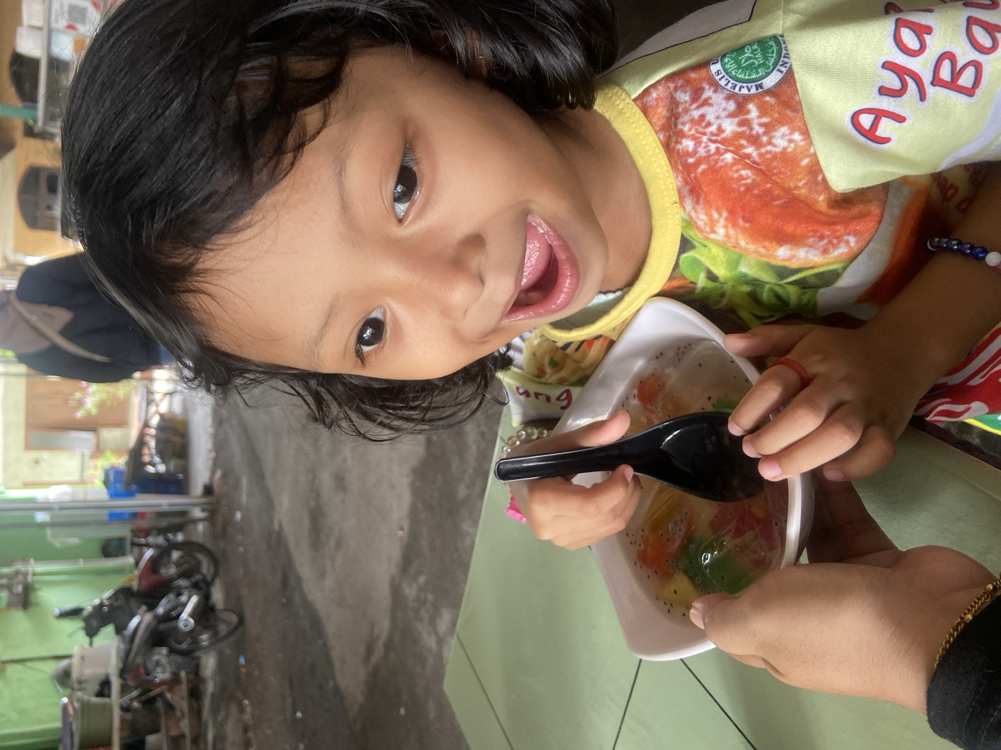 A young child with short, dark hair and brown skin holds a bowl of what appears to be fruit salad or a similar dessert. The child is wearing a yellow and orange t-shirt with writing on it. The bowl is white and contains colorful pieces of fruit-like substance in a clear liquid. A black spoon is partially submerged in the bowl. A person's hand with a gold bracelet is partially visible in the bottom right corner, seemingly holding or supporting the bowl. The scene is set on a green surface, likely a table. In the background, out of focus, is what appears to be a motorcycle and part of an urban environment. The location is consistent with the general appearance of Gelam, Indonesia, but more specific details would be required to confirm this.