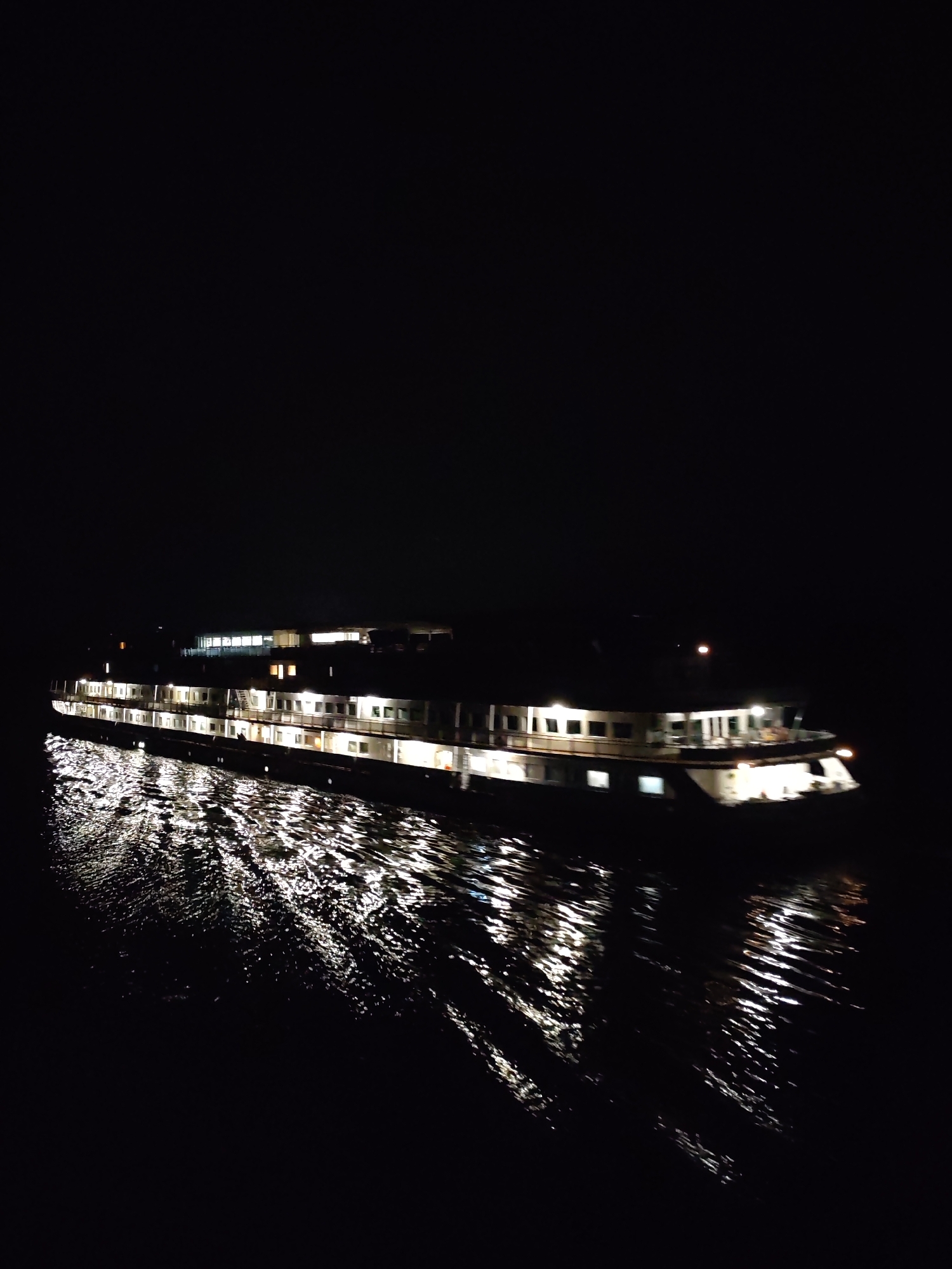 A nighttime photograph depicts a multi-level riverboat illuminated against a dark background, its lights reflecting on the water's surface. The boat appears to be moving, creating a wake visible as shimmering lines in the water. The location is unknown.