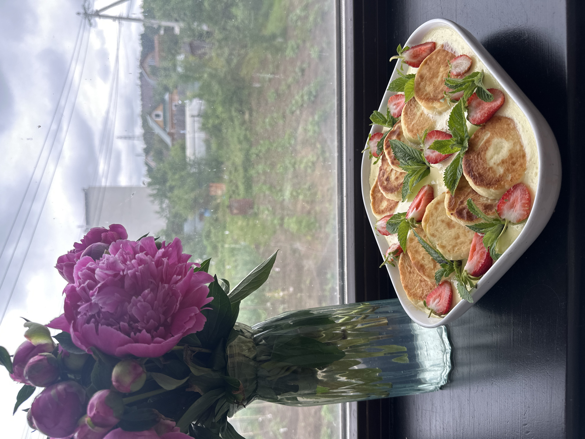 A bouquet of vibrant pink peonies in a clear glass vase sits to the left of a white oval dish filled with syrniki, which are Russian cheese pancakes. The syrniki are arranged in rows and drizzled with a creamy, pale yellow sauce. Sliced strawberries and fresh mint leaves are interspersed among the pancakes, adding pops of color and freshness. The dish is placed on a dark, flat surface, likely a windowsill. In the background, a window offers a view of a slightly overcast, rural landscape with trees, houses, power lines, and a cultivated field. The location is identified as Chaikovsky, Russia. There are no people present in the image.