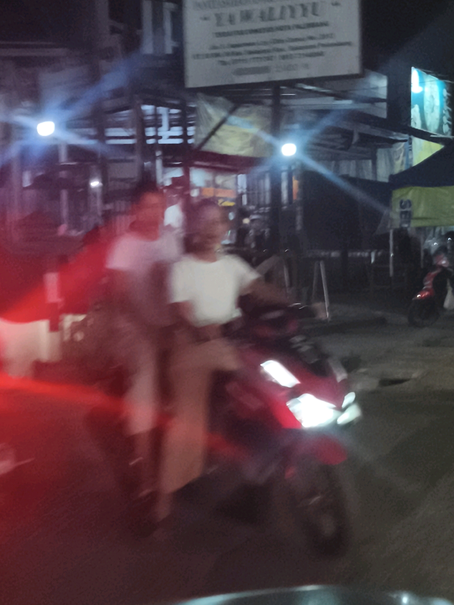 The image depicts a blurry nighttime street scene in Palembang, Indonesia. A man and a woman are riding on a red scooter. The man appears to be driving, while the woman is seated behind him. Both are wearing white shirts. The scooter's headlights are on, illuminating the road ahead. In the background, there are several structures, possibly shops or stalls, with some signage visible. A blue light from a sign is also present. The overall atmosphere is one of a busy urban environment at night, with the characteristic glow of streetlights and commercial signage. The blurriness suggests movement, likely the scooter in motion.