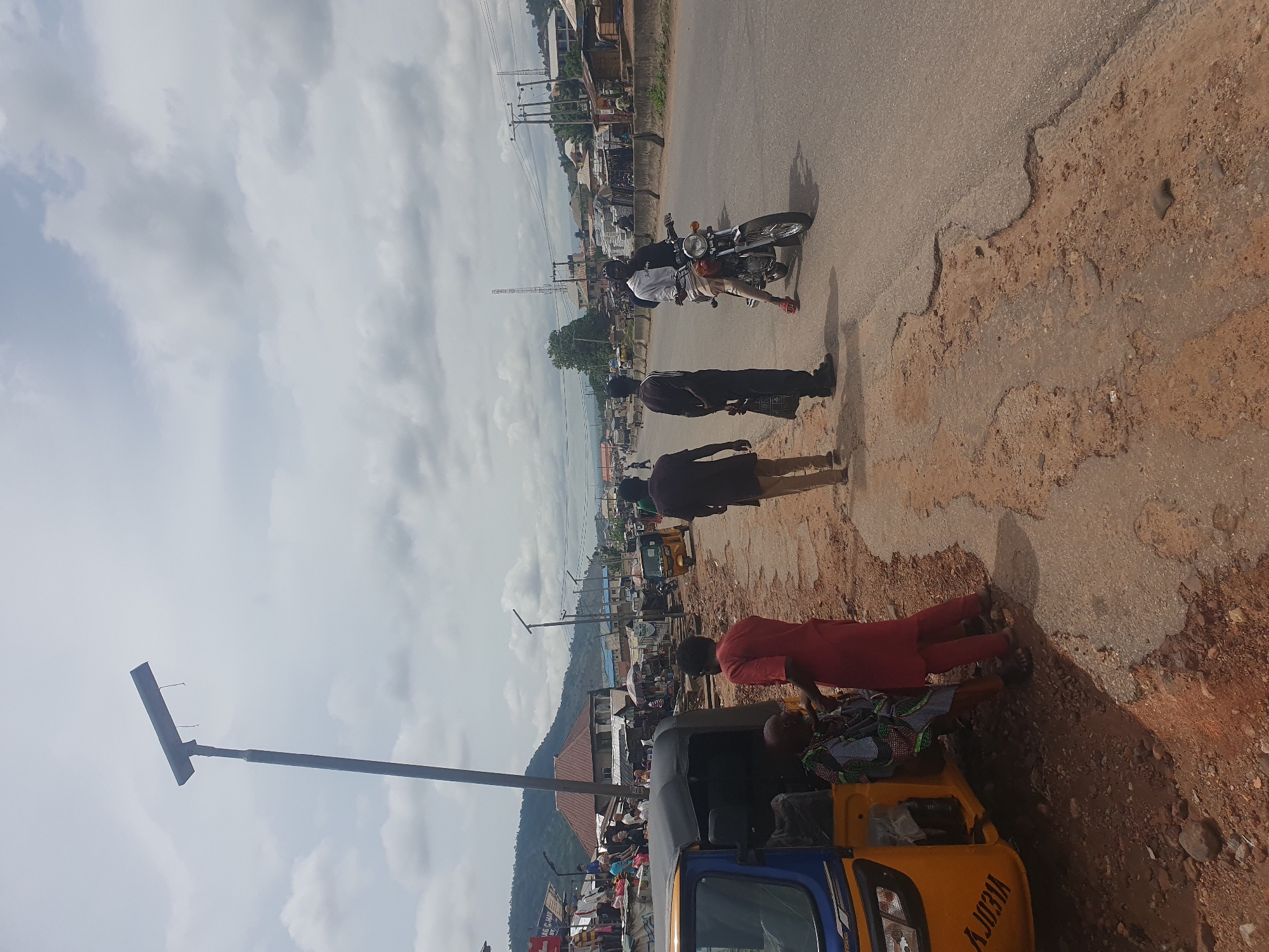 The scene depicts a street in Lokoja, Nigeria. The road is partly paved with areas of exposed earth. A motorcycle with two riders is present, along with several pedestrians. A blue and yellow tricycle (Keke Napep) is parked on the side of the road. There is an electrical pole nearby. The environment includes buildings, trees, and a sky with scattered clouds. The background suggests a hillside or mountain.