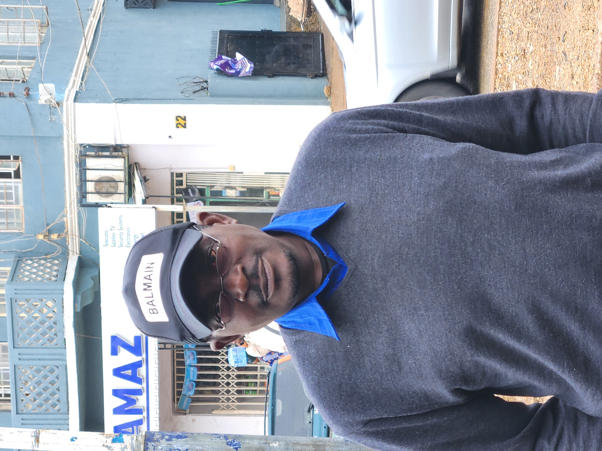 A street scene in Jos, Nigeria, featuring a man in the foreground. He is seen from the chest up, wearing a dark gray long-sleeved top over a blue collared shirt. He sports a dark baseball cap with "BALMAIN" in white capital letters across the front, and sunglasses. His expression is neutral.
The background reveals an urban environment with buildings painted light blue and white. On one building, the number "22" is visible in yellow. Below it, an air conditioning unit is mounted, and a sign partially obscured by the man's head lists services such as "Telecoms," "Satellite TV," "Security Systems," and "Electronics." Further left, a large blue sign shows "AMAZ," likely part of a business name, and smaller blue signs with "aramc" are visible in the distance. Electrical wires are strung along the buildings. A white car is partially visible on the right, parked beside a textured, light brown ground. The scene is illuminated by daylight.