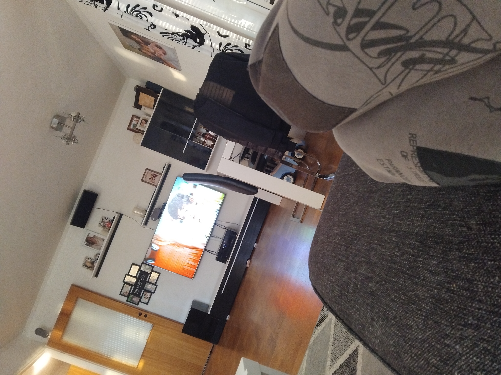 The image shows an interior view of a living room, likely in Rätan, Sweden. The room features a wooden floor and white walls. In the center of the room, a large flat-screen television is mounted on a black and white entertainment unit. Above the television, a soundbar is mounted on the wall. To the left of the television, a tall black speaker is placed on the floor. A large glass-paned wooden door is visible on the left side of the frame. The walls are adorned with several framed photographs and a black shelf holding decorative items. A modern ceiling light fixture is visible overhead. To the right, a black cabinet with glass doors displays bottles. A black jacket is draped over a chair. Curtains with a black and white pattern hang by the window on the right. In the foreground, a person is partially visible, seated on a dark-colored couch with a patterned throw pillow. The overall impression is a cozy, lived-in living space.