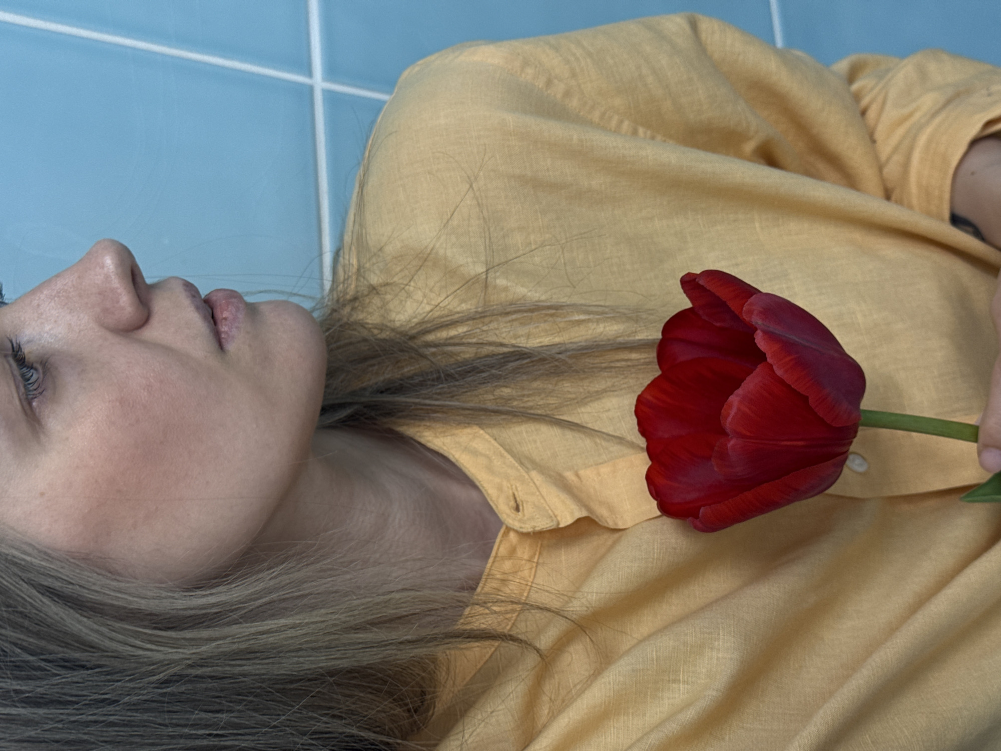 A woman with fair skin and long, light brown hair lies on her back wearing a yellow linen shirt. A single red tulip rests on her chest. The background appears to be a light blue tiled wall. There is no indication of location.