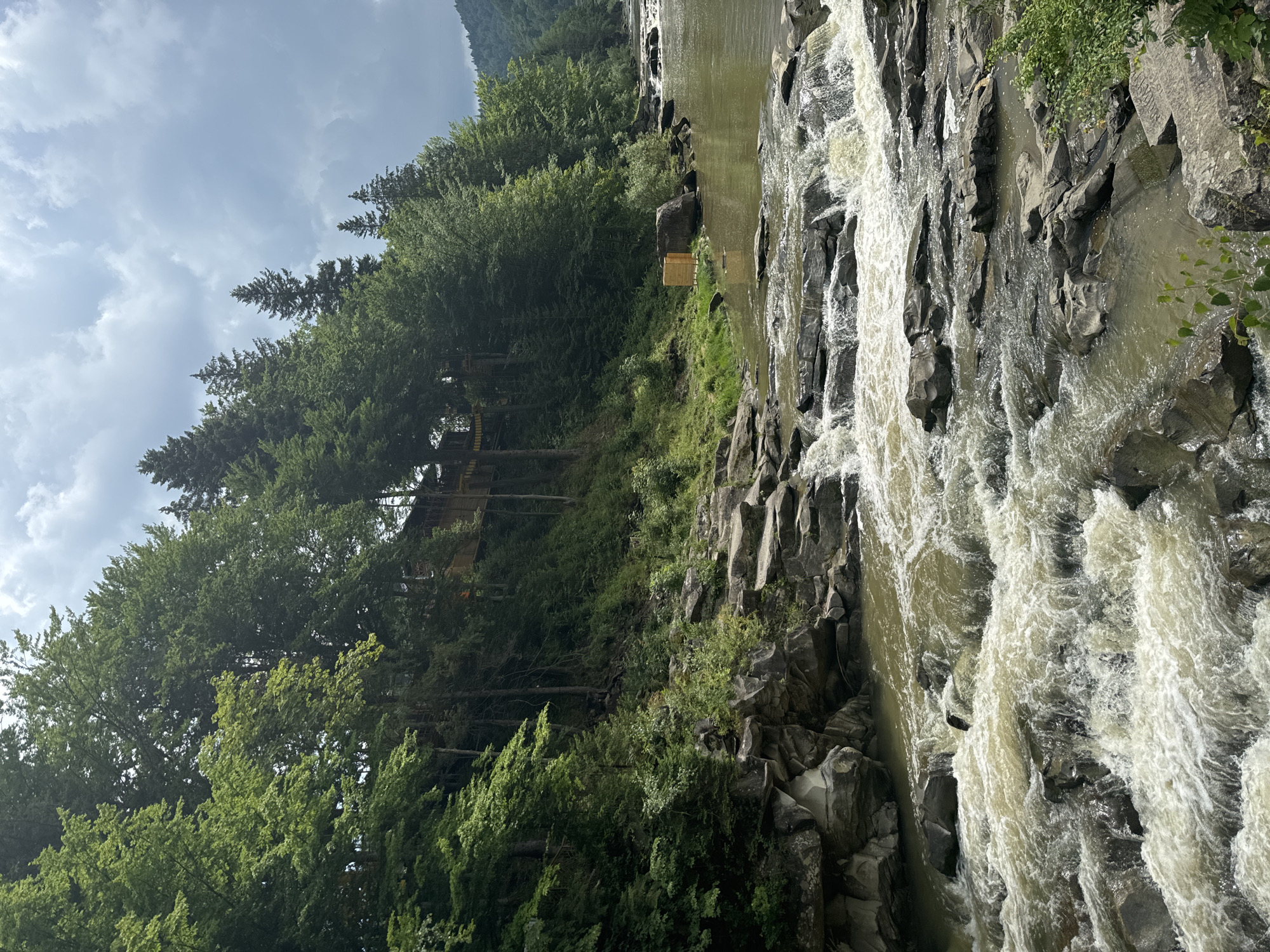This image captures a scene along the Prut River in Yaremche, Ukraine, likely near the famous Yaremche bridge and waterfall. The foreground is dominated by the turbulent, frothy waters of the river cascading over dark, jagged rocks. The water is a murky, greenish-brown color, indicative of a natural river flow.
The riverbanks are steep and heavily wooded with a dense growth of tall, green trees, primarily deciduous with some conifers interspersed. The foliage is lush and vibrant, suggesting a summer or late spring setting. On the left bank, a section of what appears to be an elevated wooden structure, possibly a tourist attraction or accommodation, is visible nestled amongst the trees on the hillside. The structure is yellow and brown, with visible railings and supports.
Further up the slope, the trees continue to cover the terrain, leading up towards a slightly overcast sky. The clouds are a mix of grey and white, hinting at the possibility of rain. On the right bank, a small, light-colored wooden structure, resembling a changing room or small shelter, is situated near the water's edge, next to a large boulder. The overall environment is natural and wild, with the river and forest being the primary elements. No people are visible in the image.