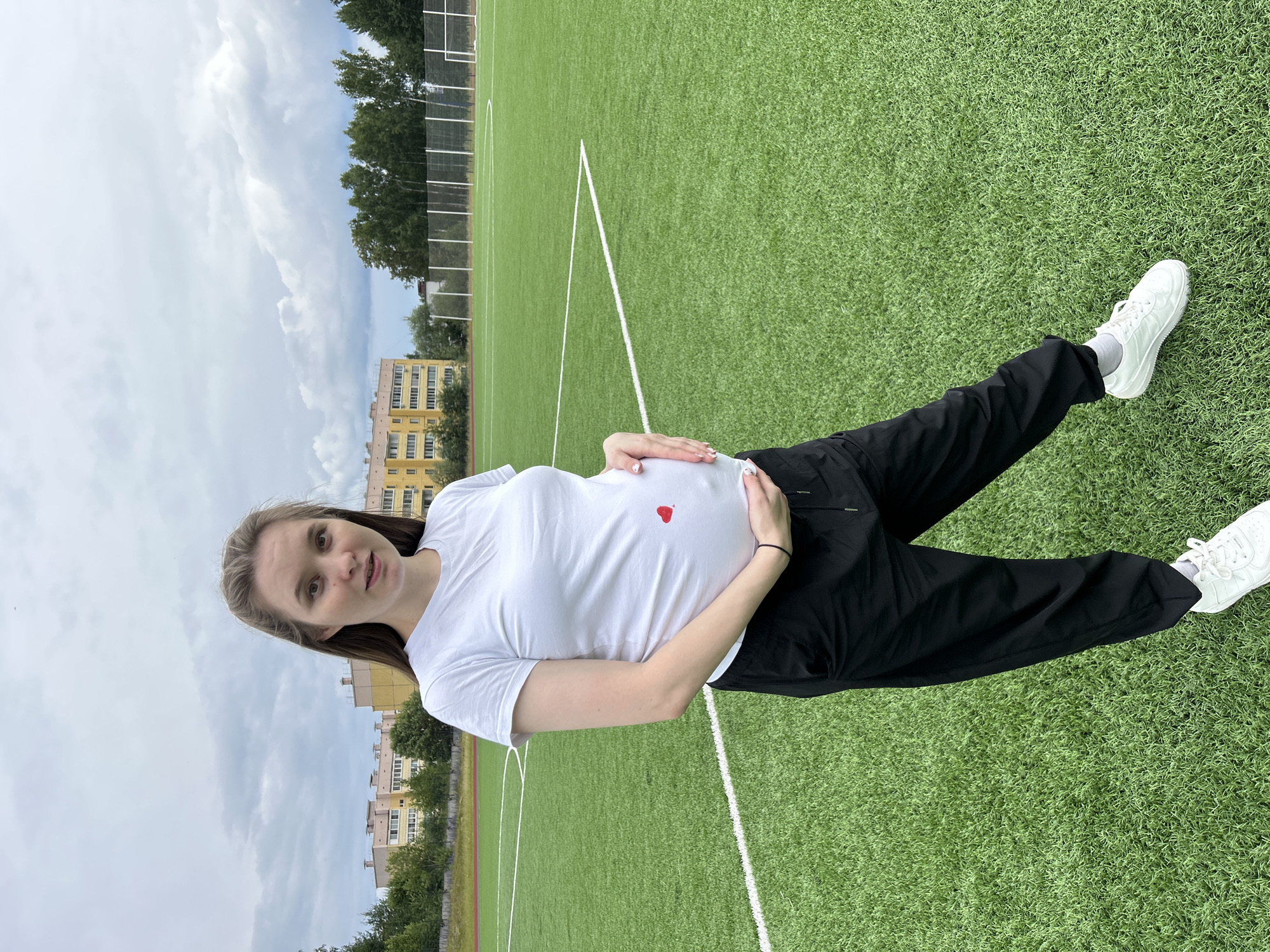 A young woman, appearing to be in her late teens or early twenties, stands on an outdoor artificial turf field, likely a sports field due to the presence of white lines. She is wearing a white t-shirt with a small red heart printed on the chest, black pants, and white sneakers. Her hair is long and light brown, and she is looking towards the camera with a slight smile. She has her hands resting on her pregnant belly. In the background, there are trees, a multi-story yellow building, and a cloudy sky. The lighting suggests it is daytime. The location is identified as Noviy, Russia.