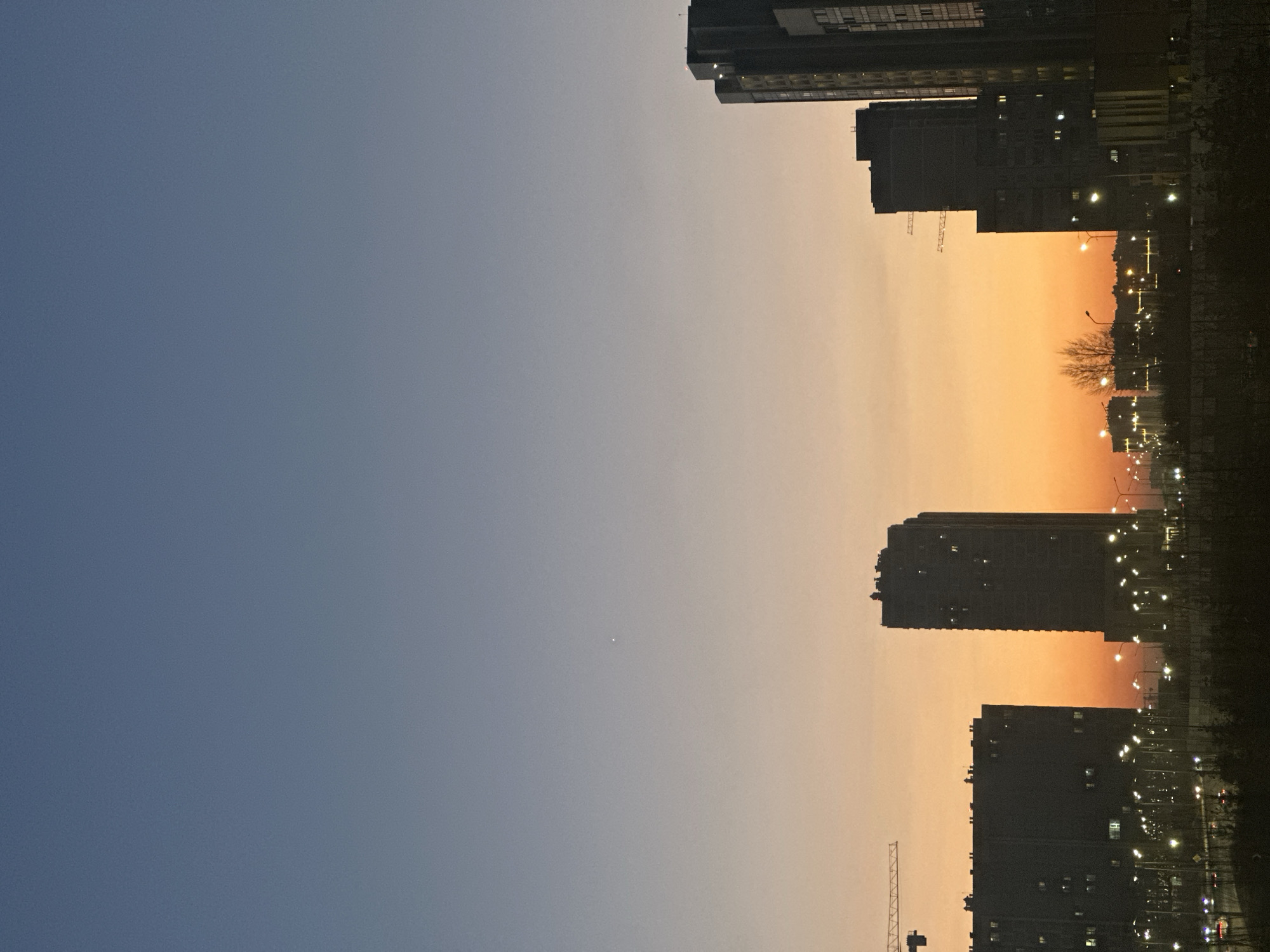 The image depicts a cityscape at dusk in Kazan, Russia. The sky transitions from blue to orange. High-rise buildings flank the right side of the frame, silhouetted against the sunset. Streetlights illuminate the road and surrounding area. A crane is visible near the bottom of the frame. There are no people visible.