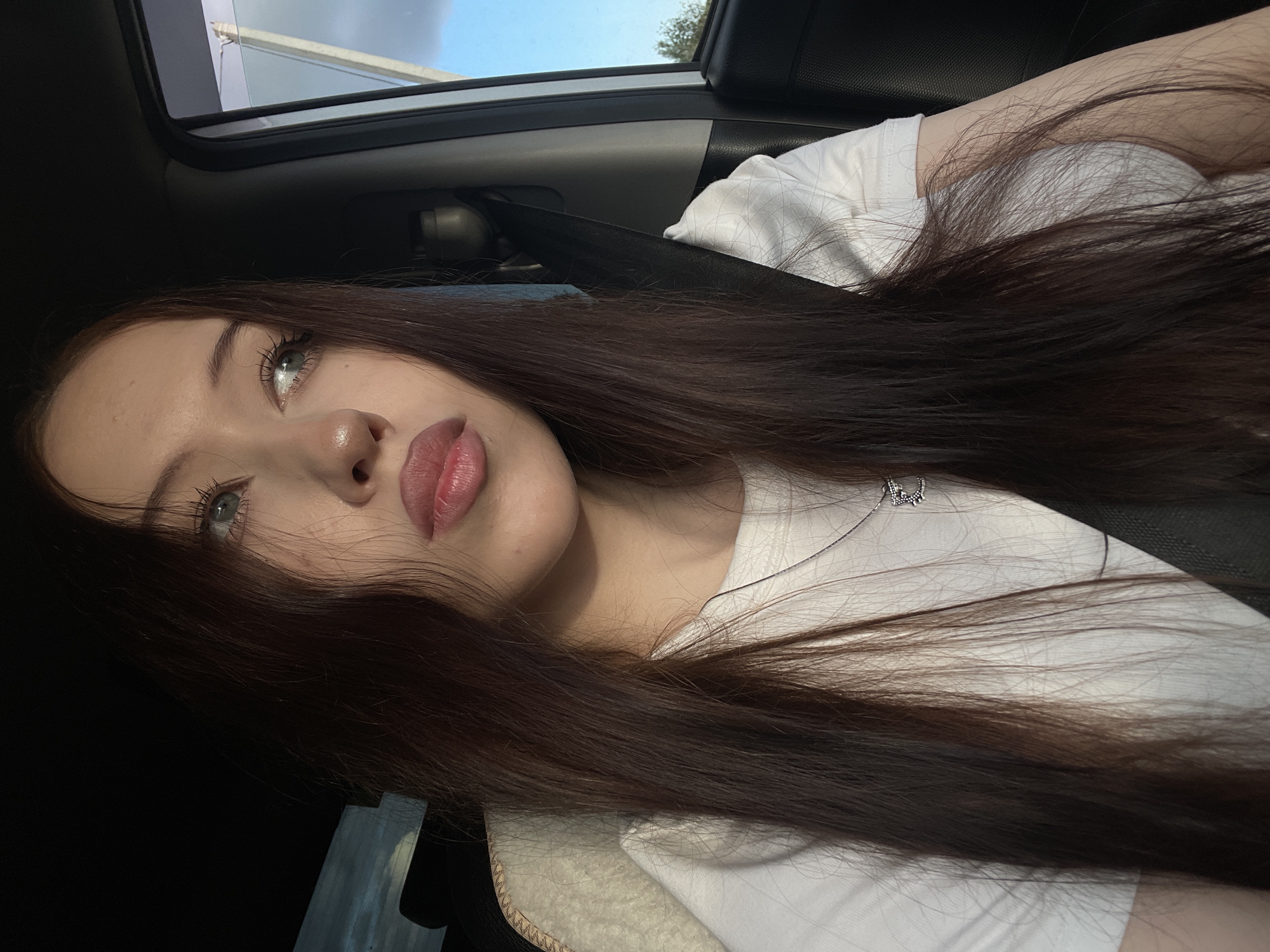 A young woman with long, dark brown hair is in the passenger seat of a car, looking out the window. She is wearing a white t-shirt and a silver necklace with a charm. The sun is shining, casting shadows in the car. The background shows a cloudy sky and some trees. The location is the city of Gavrilovka, Russia.