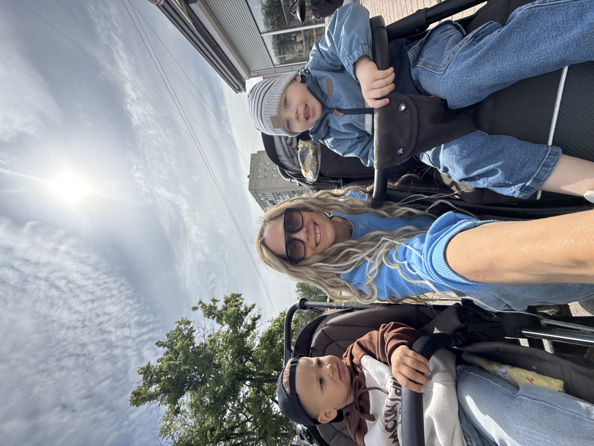 A woman with blonde dreadlocks and sunglasses is positioned between two children in strollers on a city street in Заволжье, Russia. The woman is smiling at the camera. One child wears a striped hat and a blue jacket, while the other wears a baseball cap and a brown and white jacket. A tree and a building are visible in the background. The sky is partly cloudy.