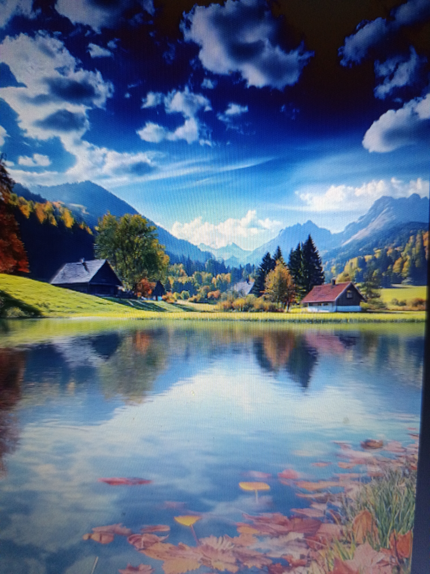 The image depicts a serene alpine landscape during autumn. In the foreground, a calm body of water reflects the sky and surrounding scenery. Scattered on the water's surface are fallen leaves, some in shades of red and yellow. Beyond the water, a gently sloping grassy hill leads up to a cluster of buildings.
On the left side of the hill, a dark-colored, rustic house with a steeply pitched roof sits nestled amongst trees displaying vibrant autumn foliage. Further to the right, a second building with a reddish-brown roof is visible, set against a backdrop of dense evergreen trees and mountains. The mountains in the distance are tinged with the warm colors of autumn, with patches of green and yellow from trees. The sky above is a brilliant blue, punctuated by dramatic, dark and light-colored clouds that create a sense of depth and movement.
The scene evokes a peaceful, natural environment, possibly a rural or semi-rural setting. It is not possible to determine the specific location within the city of Košice, Slovakia, as the image portrays a generic mountain landscape that could be found in various regions with similar topography and climate. There are no people or animals visible in the image, and the focus is on the natural beauty and the quaint structures.