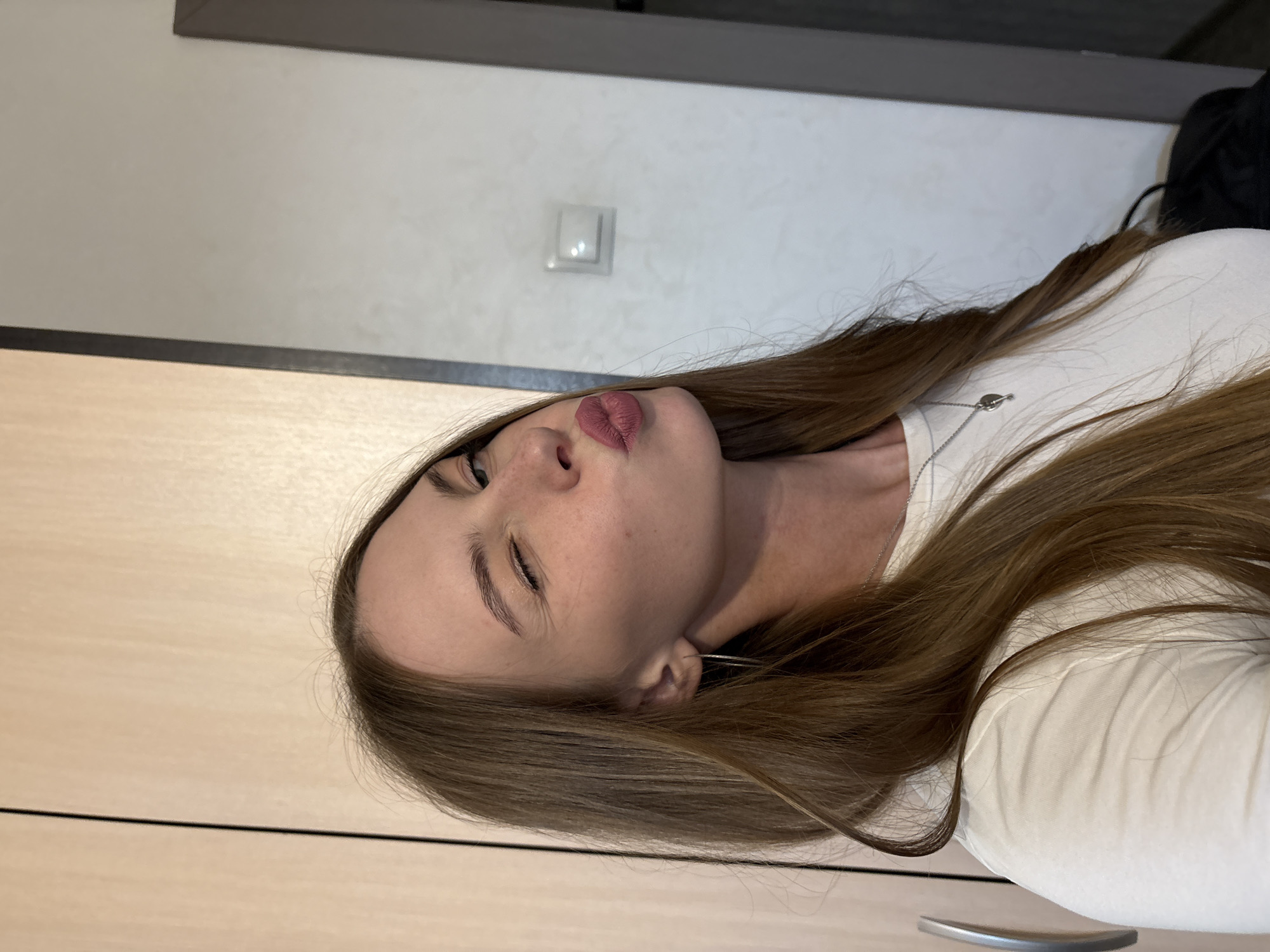 A young woman with long, light brown hair is captured in a close-up selfie. She is winking with her left eye and puckering her lips, which are coated in a matte pink lipstick. She is wearing a simple white t-shirt and a delicate silver necklace with a small, heart-shaped pendant. Her left ear, adorned with a thin hoop earring, is visible. The background appears to be an indoor setting, with a light-colored wall and a closet door with a silver handle on the left side. There is also a light switch visible on the wall. The lighting is soft and diffused. The context of the image does not indicate it is located in the city of Perm, Russia.