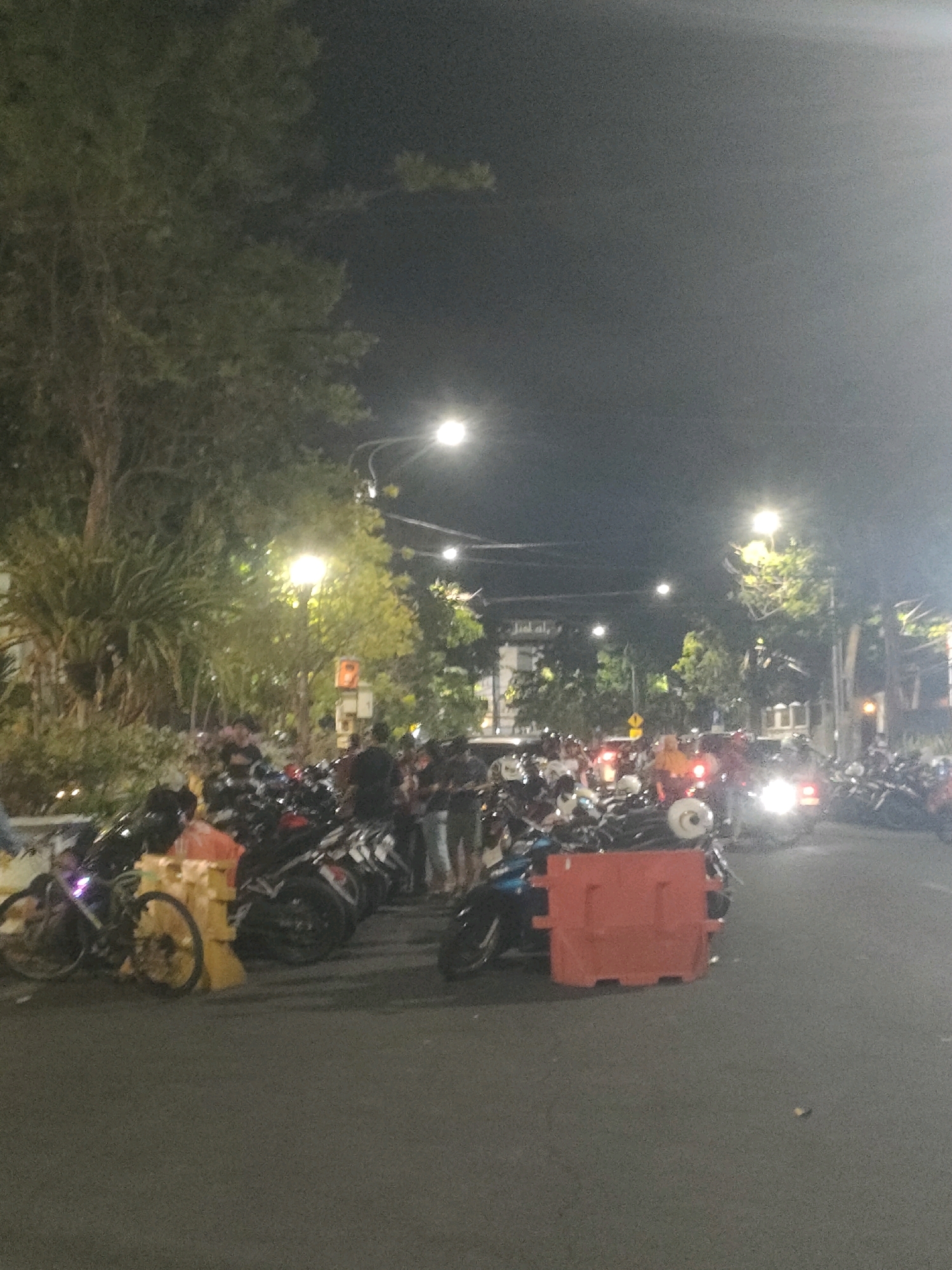 This is a nighttime, vertical, low-resolution, slightly grainy image captured on a street in Surabaya, Indonesia. The scene depicts a gathering of people and numerous parked motorcycles, suggesting a social event, a market, or a parking area.
In the foreground, a dark asphalt road is visible. To the left, a bicycle with a neon light on its wheel is parked. Behind it, a row of motorcycles is densely packed. Some of these motorcycles are black, while others have red and white accents. There are also some orange and yellow barriers placed in front of some of the motorcycles.
Further into the background, more motorcycles are visible, and a crowd of people is gathered. The people are standing and appear to be socializing or waiting. They are dressed in casual attire.
The environment includes trees with green foliage on the left side of the image, some of which are illuminated by streetlights. Several streetlights are also visible along the road, casting a yellowish glow. Buildings and structures are dimly visible in the background, suggesting an urban setting. The overall atmosphere is lively, with a sense of community activity taking place after dark.