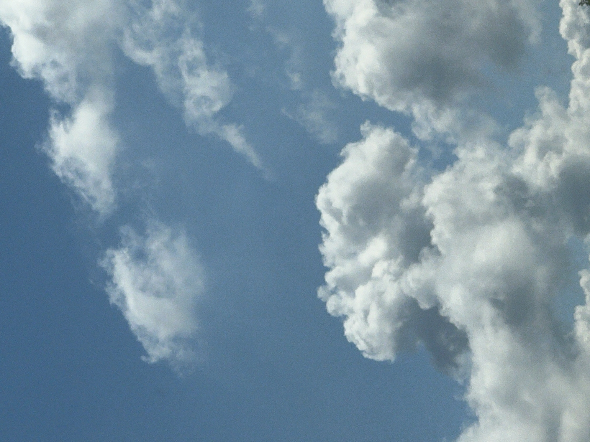 The image shows a clear blue sky with scattered white cumulus clouds. The clouds vary in size and density, with some appearing fluffy and bright, while others are more diffused and grayish. The lighting suggests it is daytime, likely sunny. There is no indication of any specific location within Warsaw, Poland, or any identifiable subjects, objects, actions, or interactions. The scene is a natural, outdoor environment focused solely on the sky and clouds.