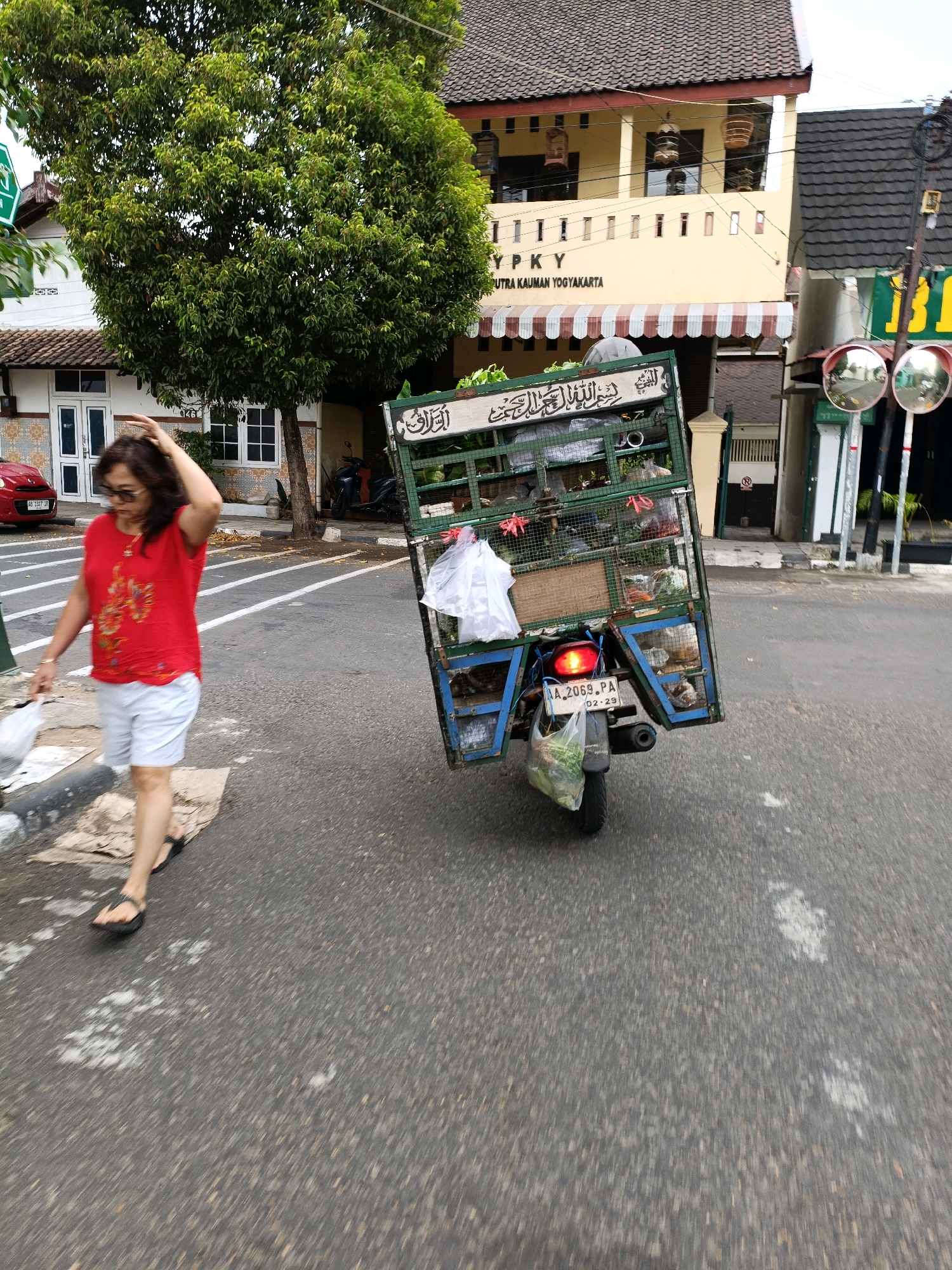 The image shows a street scene in Yogyakarta, Indonesia. In the foreground, a woman in a red shirt and white shorts walks on the asphalt road, holding a plastic bag in her left hand and touching her hair with her right hand. Behind her, a modified motorcycle with a large cargo cage attached is moving away from the camera. The cage is filled with various items, including what appears to be produce, and has a license plate that reads "AA 2069 PA". Buildings line the street, with one prominent building featuring a yellow facade and a sign that reads "YPKY" and "PUTRA KAUMAN YOGYAKARTA". Red cars are parked on the side of the road, and a large tree with dense green foliage stands near the buildings. Convex mirrors are mounted on poles along the street. The scene is captured in daylight.
