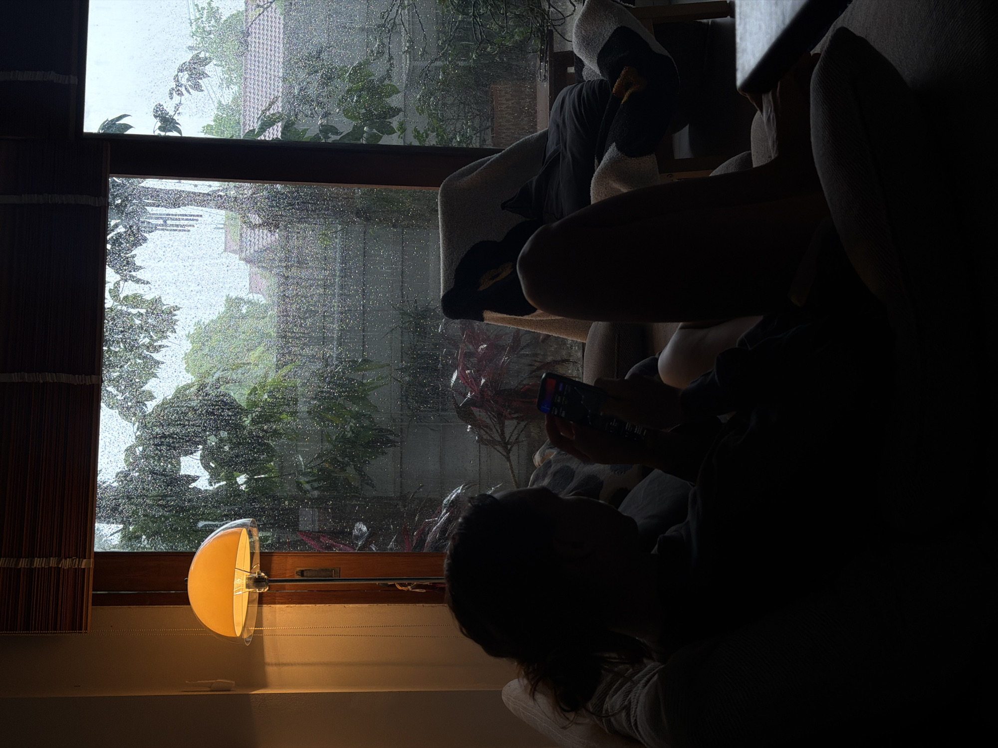 The image depicts an indoor scene at dusk, with rain visible through a large window. In the foreground, a person is silhouetted on a couch, holding a smartphone and appearing to be engaged with it. To the right, another person is reclined in an armchair, also silhouetted and partially covered by a blanket. The window reveals a rainy atmosphere with trees and rooftops of houses visible outside, suggesting a residential area in the city of Chang Phueak, Thailand. A floor lamp with a warm, orange glow is positioned to the left of the window, illuminating the interior and casting shadows. The overall mood is cozy and introspective, characteristic of a rainy day spent indoors.