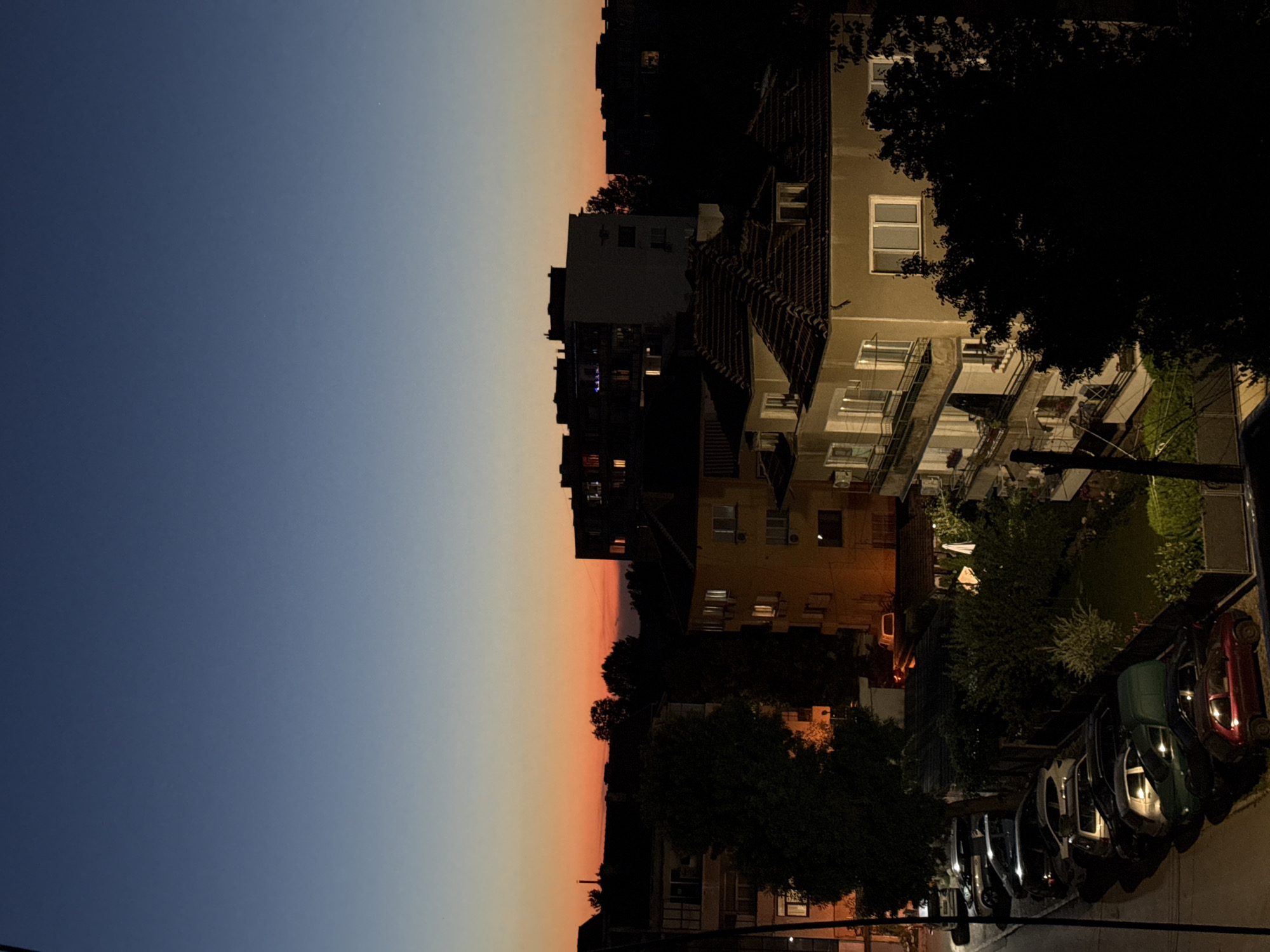 An aerial shot captures a twilight scene in Vratsa, Bulgaria. The sky transitions from a deep blue at the top to a vibrant orange and red near the horizon, indicating the sun has recently set. Silhouetted against the colorful sky are the roofs and upper floors of several buildings, including apartment blocks and a smaller, tiled-roof house in the foreground.
The foreground is dominated by a row of parked cars on a street or parking area. These vehicles are a mix of sedans and SUVs, with varying colors including red, dark green, white, and silver. Some of the buildings have lights on, casting a warm glow from their windows. Trees, some dark and full of leaves, frame parts of the scene, adding to the evening atmosphere. A utility pole is visible on the left side of the frame. The overall impression is a quiet, residential neighborhood as day turns into night.