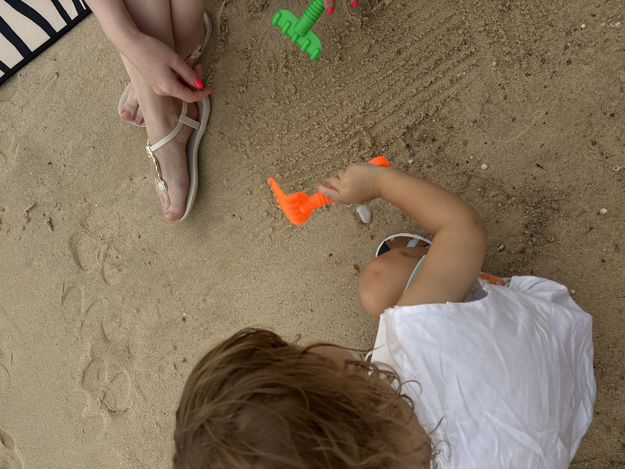 A high-angle shot shows a child and an adult's legs on a sandy beach. The child, facing away from the camera, has brown hair and is wearing a white t-shirt and sandals. They are sitting on the sand and holding an orange toy rake. To the right, an adult's legs are visible, wearing white sandals with gold embellishments. Their fingernails are painted neon pink. A black and white striped object, possibly a towel or bag, is partially visible in the upper right corner. The sand has small pebbles and shell fragments scattered across it. Footprints are visible in the sand in the background. The scene appears to be on a beach in Choeng Thale, Thailand.