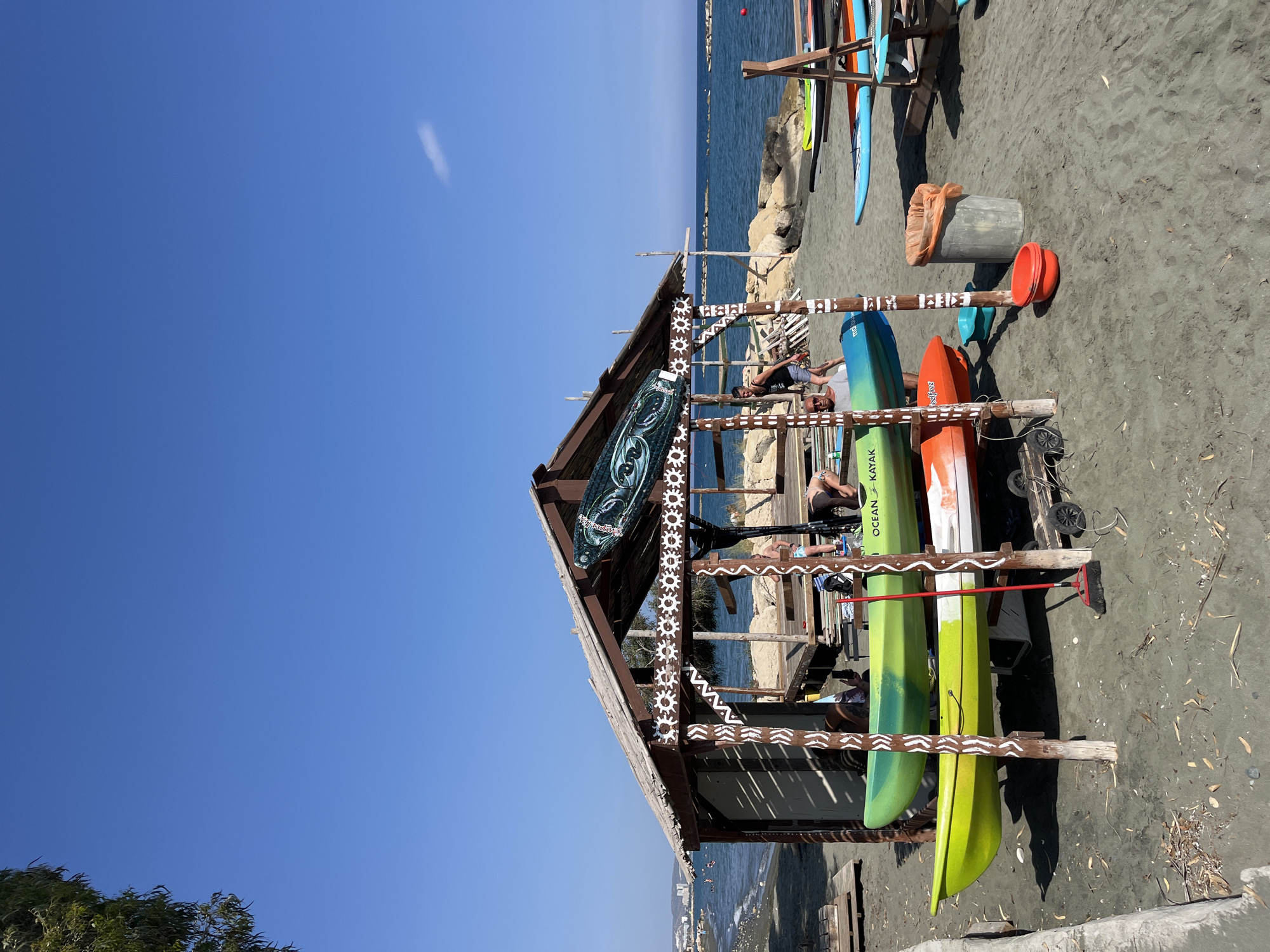 The image captures a sunny scene at the beach in Germasogeia Municipality, Cyprus. In the foreground, there is a wooden rack structure used to store colorful kayaks, including "Ocean Kayak" models. The rack is adorned with white geometric patterns. Several people are lounging inside the rack's shaded area. Adjacent to the rack are additional kayaks and paddleboards stored on the sandy beach. In the background, the blue sea extends towards the horizon under a clear blue sky. A few buildings are visible in the far distance.