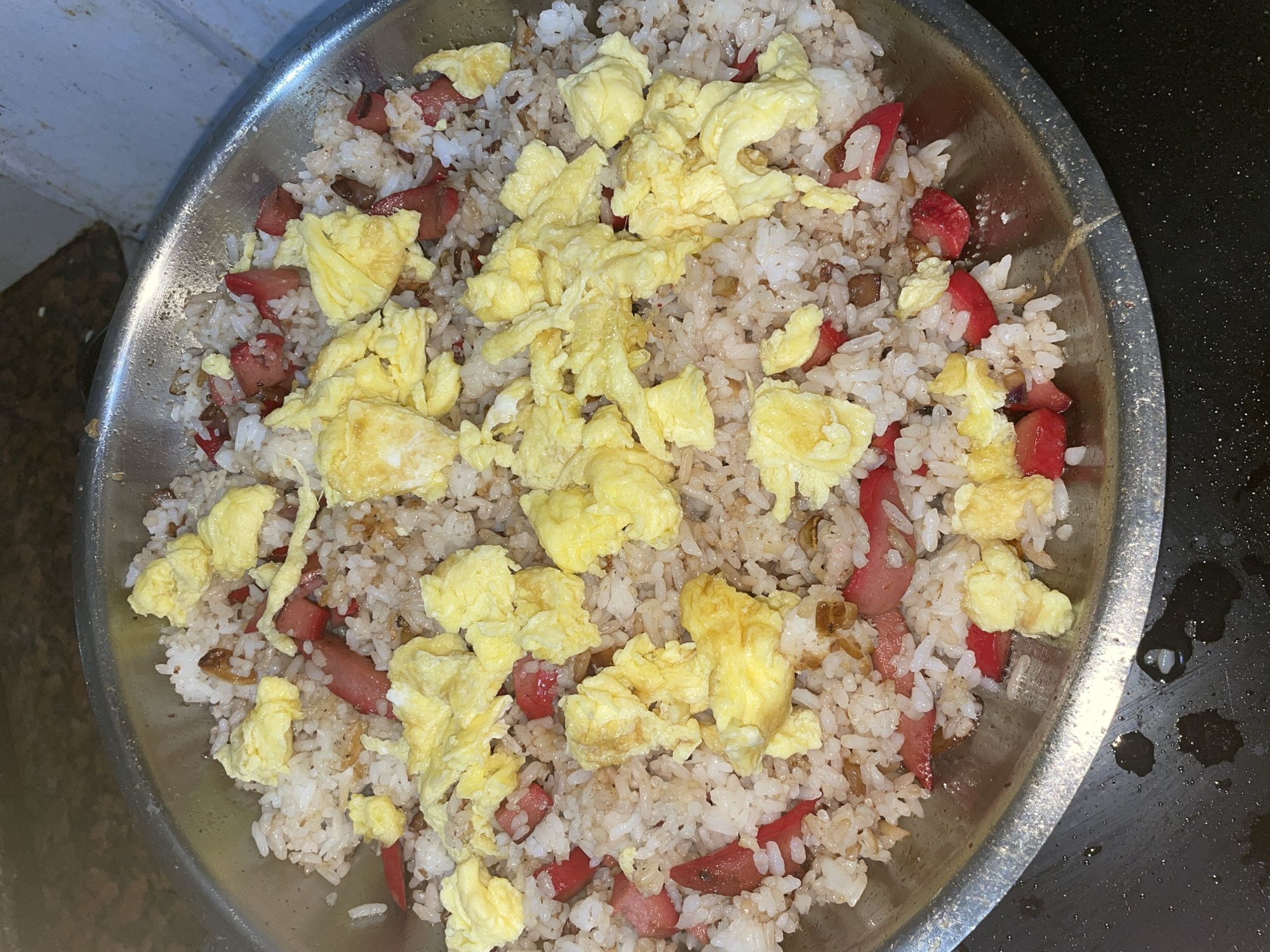 A close-up, overhead shot shows a large, metal bowl filled with Filipino-style fried rice, also known as "Sinangag." The rice is a light brown color, indicating it has been seasoned, and is mixed with visible pieces of chopped red hot dogs and scrambled eggs. The eggs are a bright yellow and are scattered generously over the top of the rice. Some darker, smaller pieces, possibly garlic or other seasonings, are also mixed in. The bowl is sitting on a dark surface, with some water droplets visible on the surface to the right of the bowl. The lighting appears to be artificial, suggesting the photo was taken indoors. There are no people visible in the image. The background to the left is out of focus but appears to be a white wall with a darker, textured area below it. This image likely depicts a homemade meal, typical of Filipino cuisine, perhaps prepared in a kitchen setting in Davao City, Philippines.