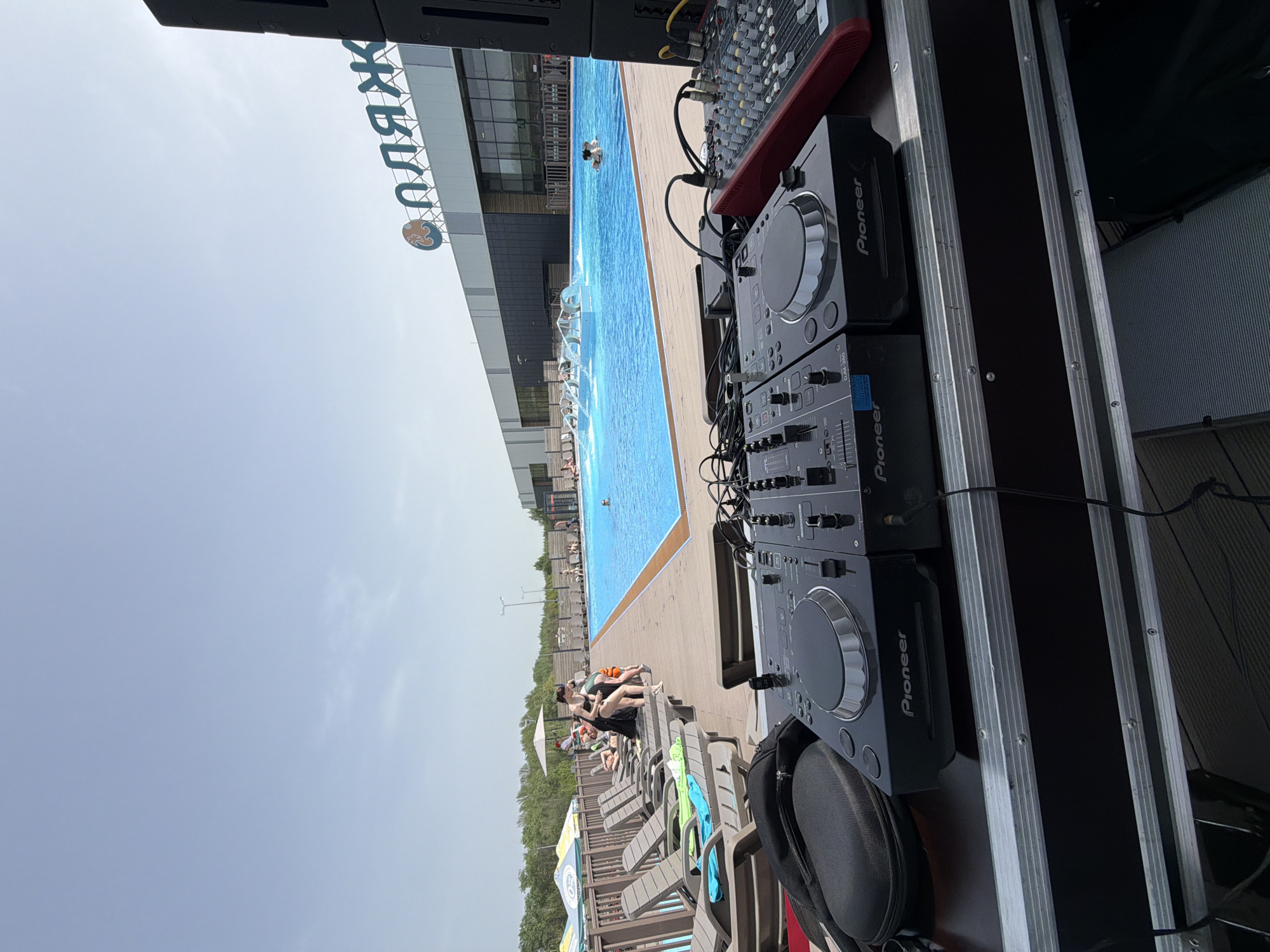 The image shows a poolside setting in Kazan, Russia. A DJ setup with two Pioneer CDJ-350 units and a red audio mixer is positioned next to a pool. The building in the background has the word "ПЛЯЖ" (which means "beach" in Russian) on its facade. People are swimming in the pool and relaxing on lounge chairs nearby. The sky is overcast. The camera is positioned close to the DJ equipment, giving a view of the pool area and the building.