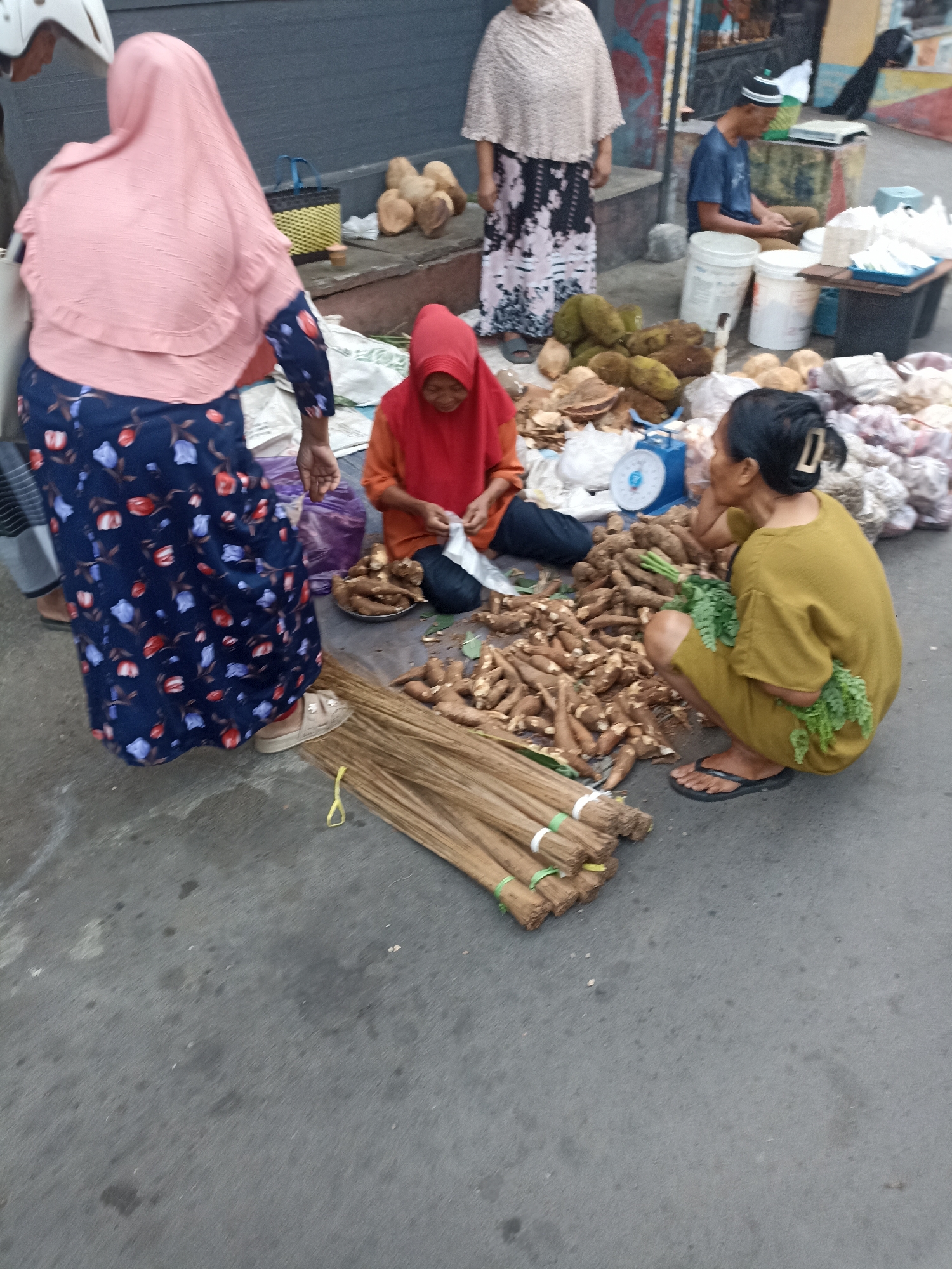 The image depicts an outdoor market scene in Mojopanggung, Indonesia. In the foreground, a woman in a yellow outfit is squatting and holding a bunch of leafy greens, with a pile of root vegetables in front of her. Next to her is a stack of brooms. To her left, another woman in a red headscarf is seated and peeling what appear to be root vegetables. A woman in a pink headscarf and a floral dress stands to the left, partially obscuring another person. In the background, several other individuals are visible, along with market stalls displaying produce and other goods. The ground is paved with asphalt, and there are various containers and bags scattered around. The overall impression is of a busy marketplace where fresh produce is being sold.