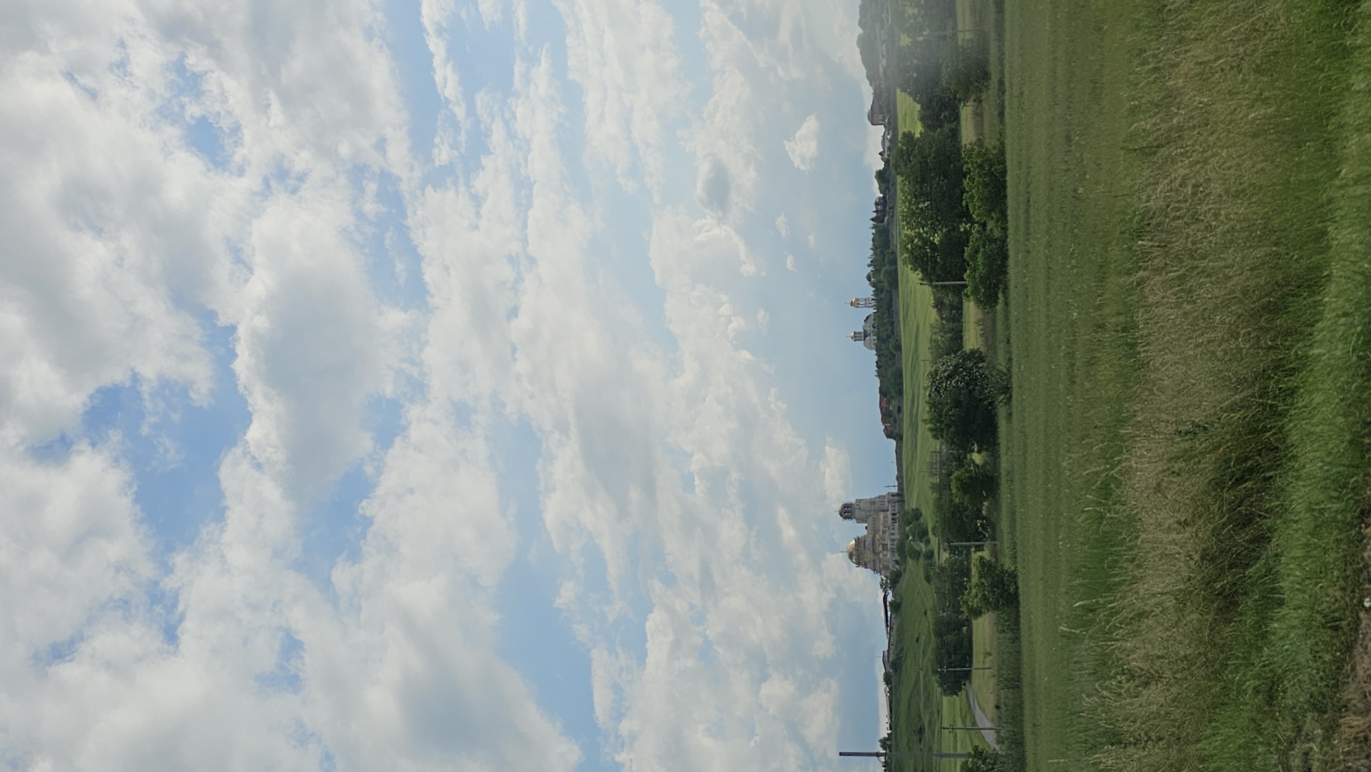 The image depicts a pastoral scene with a wide expanse of green grass in the foreground, stretching towards a distant hillside dotted with trees and buildings. The sky is a prominent feature, filled with fluffy white clouds against a backdrop of pale blue.
The scene appears to be set in Yessentuki, Russia, given the location context. The buildings on the hillside, particularly the one with golden domes, suggest a possible religious or historical site. The overall atmosphere is serene and peaceful, indicative of a pleasant day, likely in the late morning or early afternoon, judging by the bright, diffused light. The weather seems fair, with the clouds suggesting a partly sunny day. There are no visible people or specific activities taking place, making it a landscape-focused image. The tall grass in the foreground is dry and wispy, hinting at warmer weather, possibly late spring or summer. The composition emphasizes the vastness of the landscape, with the sky dominating the upper portion and the rolling green fields leading the eye towards the distant structures.