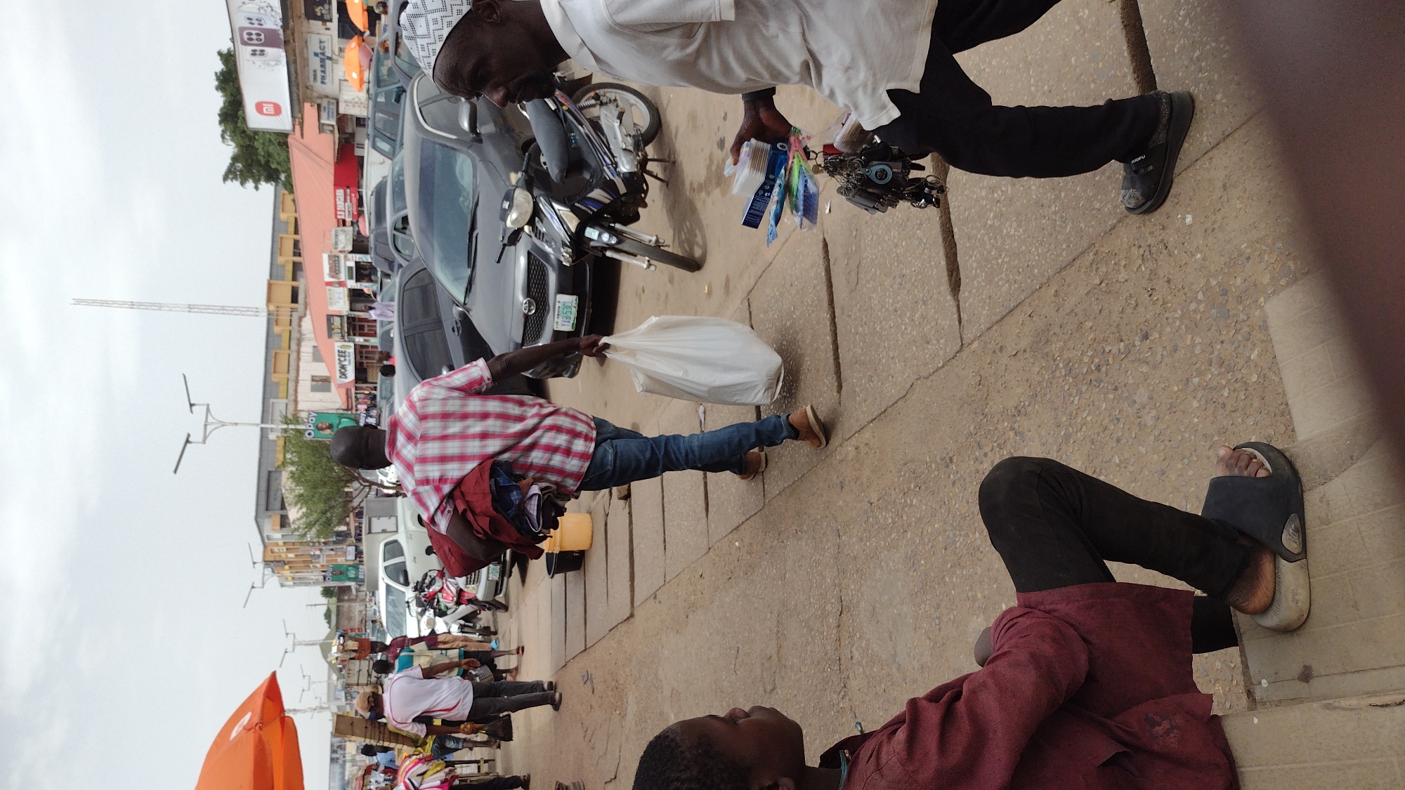 The image, taken from a low angle, captures a street scene in Jos, Nigeria. The ground is paved with concrete. A man in a red and white plaid shirt, blue jeans, and brown shoes is walking, carrying a white plastic bag in his right hand and a yellow bucket in his left. A man in a white long-sleeved shirt and black pants stands, holding several keychains. A motorcycle is parked nearby. Cars are parked along the street. Other people are walking and standing along the street, including a person wearing a red shirt and black pants sitting. Buildings, including shops, line the street. A communication tower and streetlights are visible in the background. An orange umbrella provides shade for vendors.