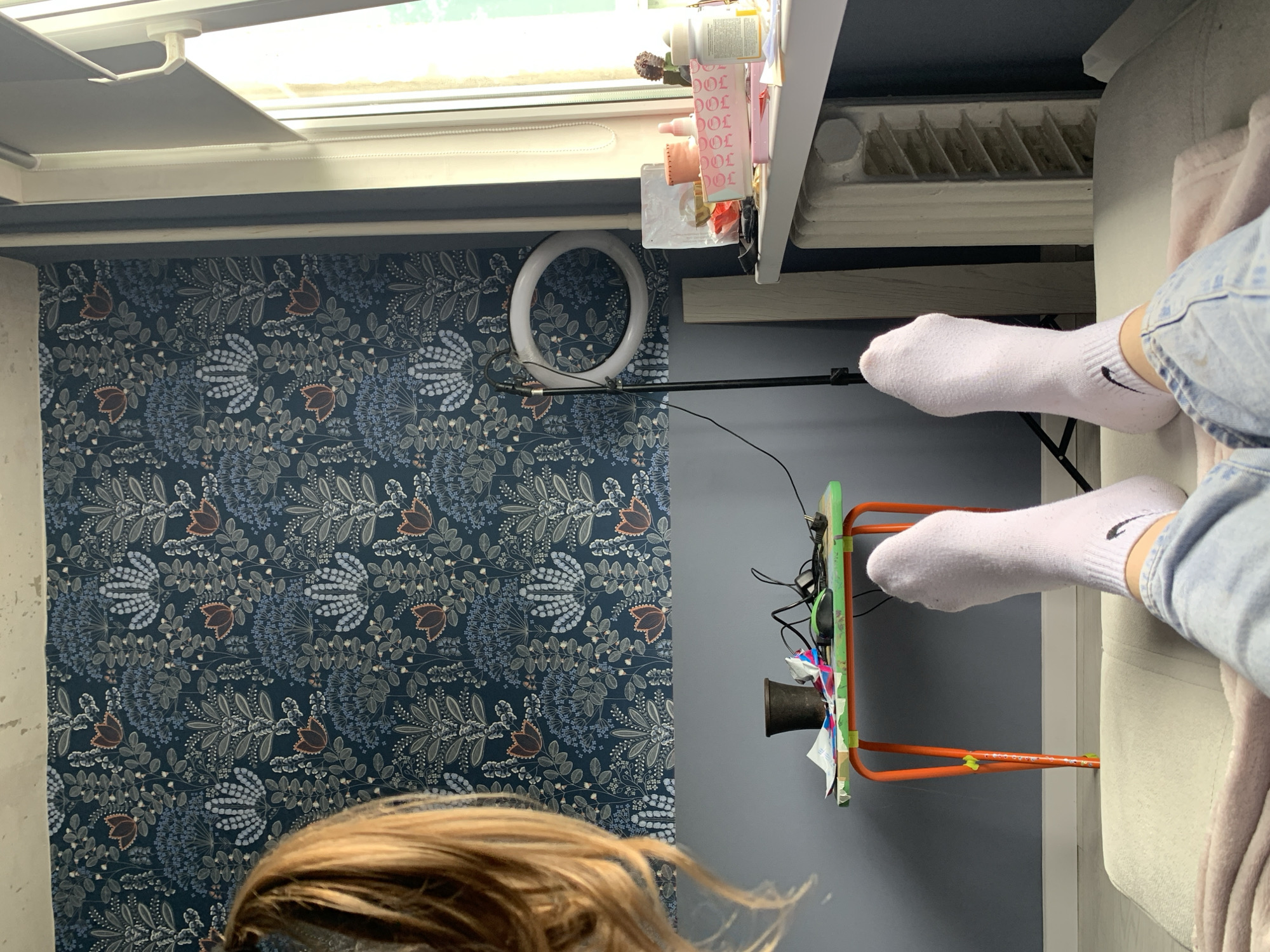 The image shows a cluttered room with a wallpapered wall featuring a dark blue floral pattern on the left. A person's hair is visible in the bottom-left corner. A ring light is mounted on the wall near the ceiling. An orange structure with a green panel is attached to the wall, and various wires and objects are hanging from it. A person's legs wearing white socks and denim jeans are visible, extending over a light-colored couch, which is positioned perpendicular to the wall. A white radiator is partially visible near the top. Bottles and other items are placed on a shelf or ledge near the top of the image. There is a skylight or window at the top. There is no visible information that indicates the location is Stavropol, Russia.