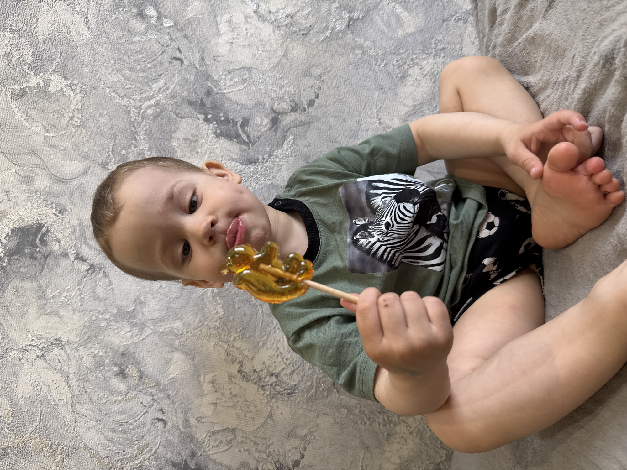 A young child, likely a toddler, is depicted in a high-angle shot, seated on a textured, light grey fabric surface, possibly a bed or sofa. The child is oriented diagonally, leaning back against a wall covered in a light grey and white patterned wallpaper with a distressed or marble-like appearance.
The child is wearing a short-sleeved olive green t-shirt with a prominent black and white print of a zebra's head on the chest. They also have on dark shorts patterned with white soccer balls. With light brown hair, the child looks directly at the viewer, mouth slightly open, tongue extended as if in the act of licking.
In their right hand, the child holds a clear, amber-colored lollipop on a small wooden stick. The lollipop is distinctly shaped like a small bird or duck. The child's legs are bent, and their left foot is visible, resting on the soft surface.
The scene takes place indoors. There are no specific visual cues within the image to indicate the provided location of Rusanovo, Russia, nor is the time of day or weather discernible. The child appears focused and engaged with the lollipop, exhibiting a relaxed and possibly playful expression. No visible text is present in the image.