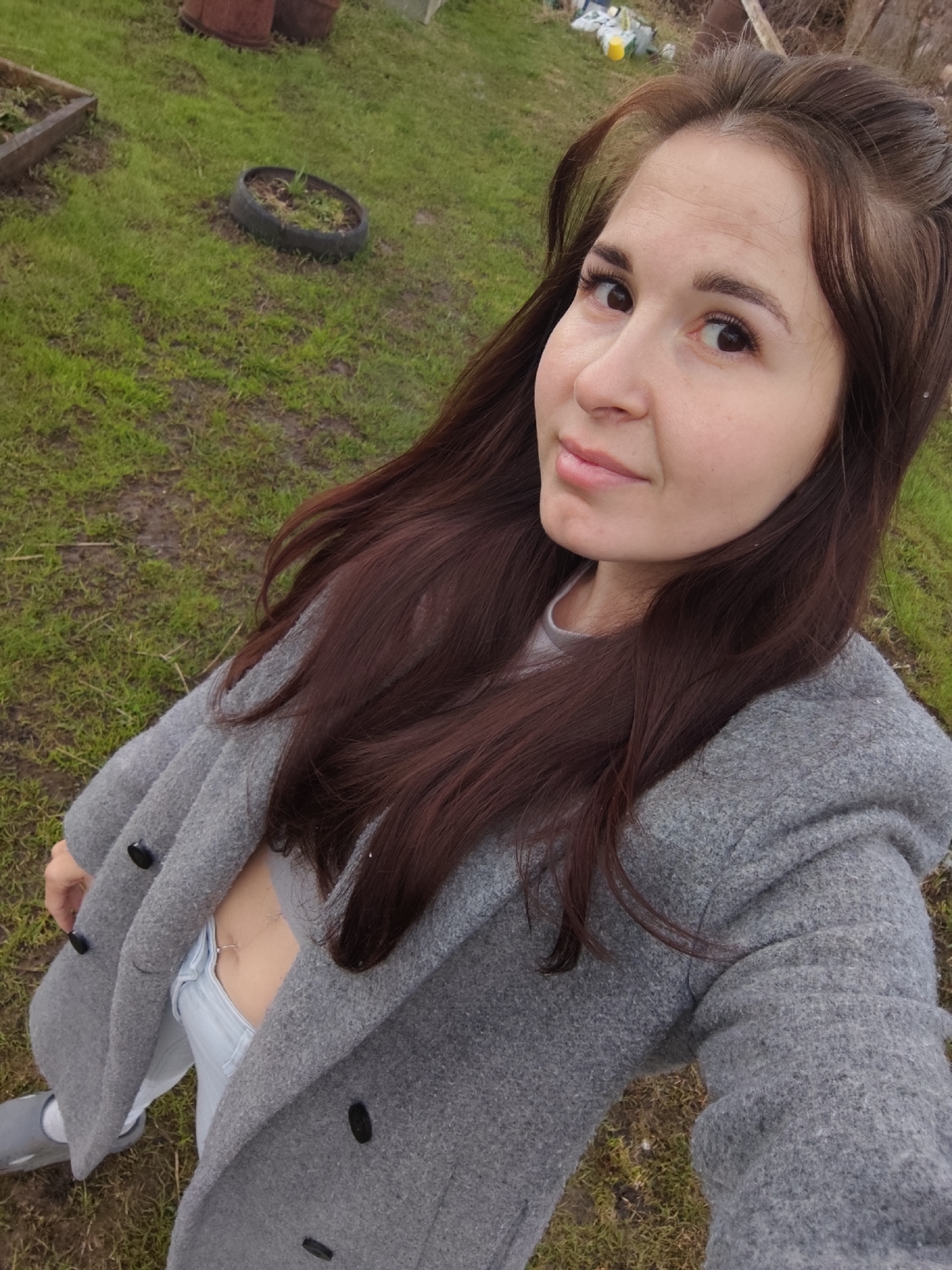 A young woman with long, dark brown hair poses for a selfie outdoors. She is wearing a light gray coat, light blue jeans, a light gray shirt, and gray shoes. The coat is unbuttoned, revealing a glimpse of her abdomen.
The setting appears to be a grassy yard or field, with a tire planter visible in the background along with other gardening implements. Based on the information provided, the location is near the city of Фоки, Russia.