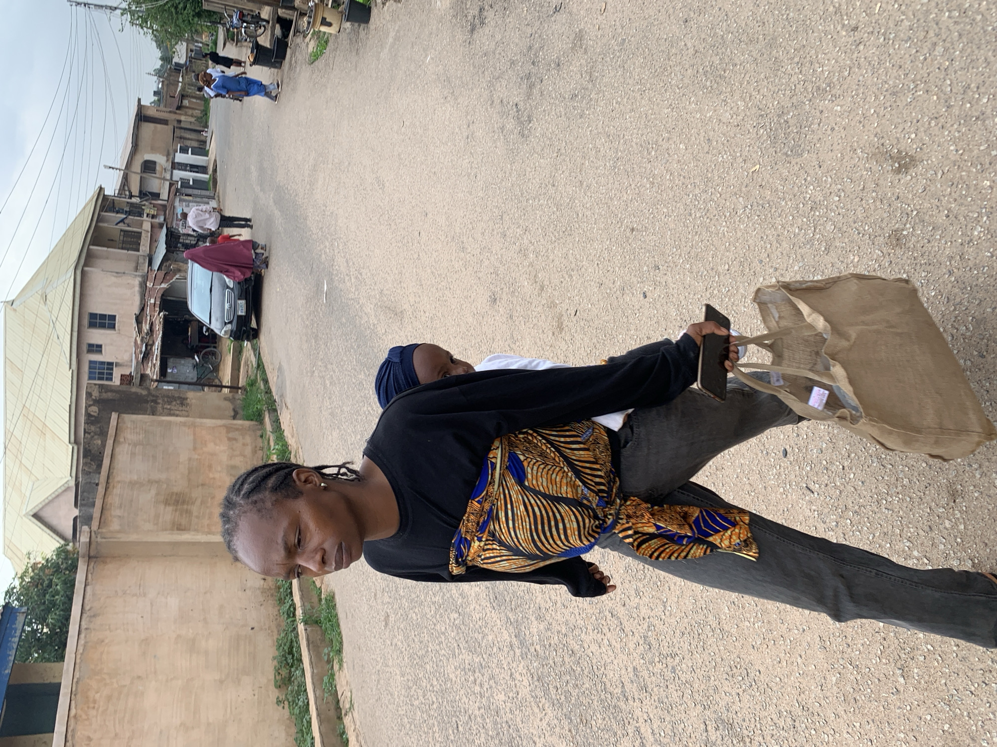 A woman in Jos, Nigeria, is walking on a paved road. She is wearing a black shirt, grey jeans, and carrying a brown tote bag. She is also carrying a child secured on her back with a blue and gold patterned cloth. She is holding a cell phone in her hand. In the background, there are buildings and other people. A dark car is parked in the middle of the road with a maroon covering, and several people are milling around the car. A person in blue scrubs can be seen further in the background.