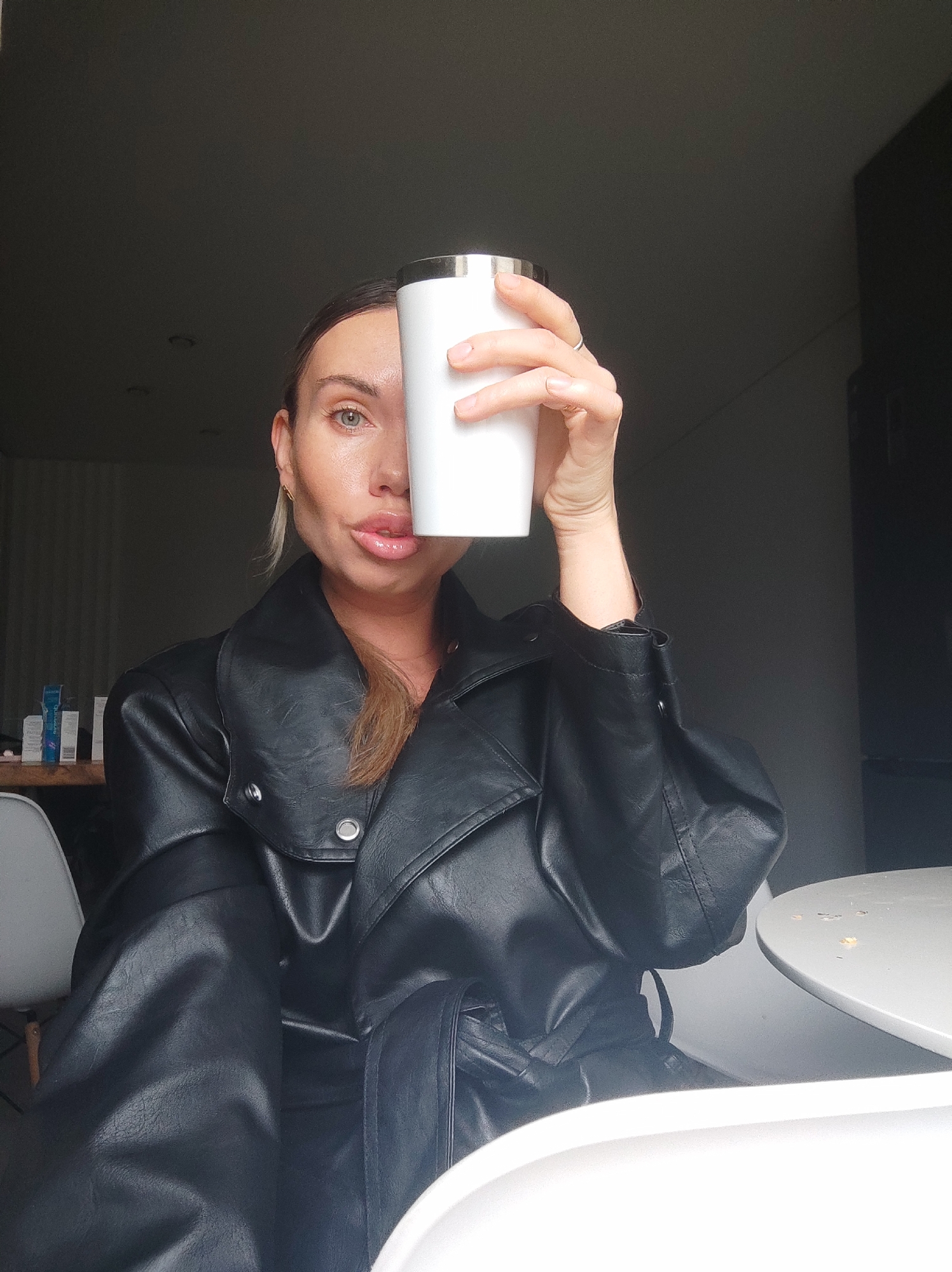 A woman in a black, belted leather jacket is holding a white tumbler in front of her face, obscuring the right side. Her left eye is visible. She has fair skin and light brown hair pulled back. She is wearing a ring on the hand holding the tumbler. The background is a dimly lit room with a table and chair in the foreground. There are some items on a surface in the background. The location is not indicated in the image, thus the location in Votkinsk, Russia, cannot be confirmed.
