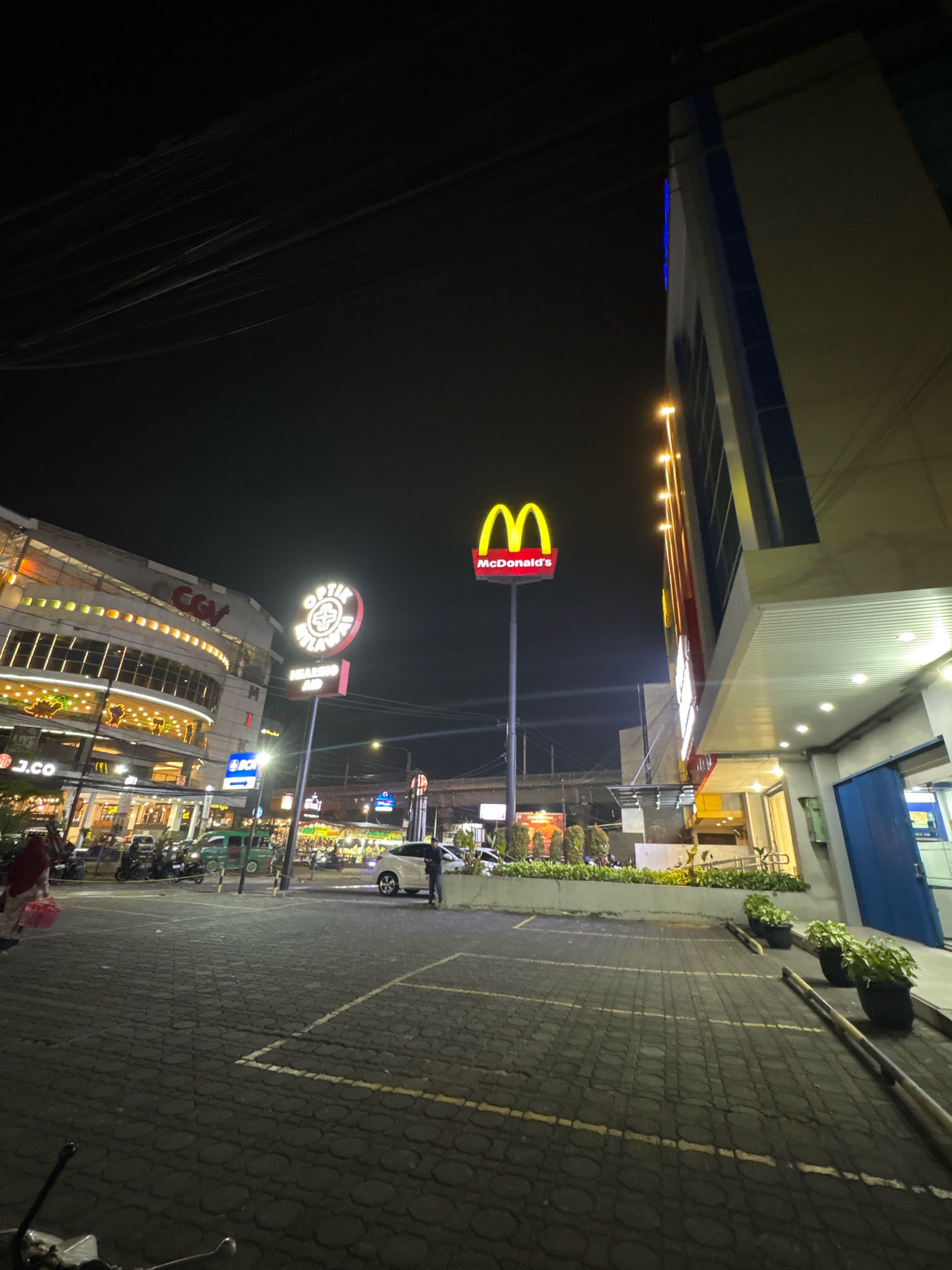 The image shows a nighttime view of a street in Bandung City, Indonesia. In the foreground, a paved parking lot with white lines is visible. Several parked motorcycles and a green car are on the left side of the image. To the right, a building with a blue facade and illuminated windows is present, with potted plants lining the walkway. Further in the background, a large multi-story building with a curved facade and glowing lights can be seen. A prominent McDonald's sign with the iconic golden arches and "McDonald's" text is mounted on a pole in the middle ground, illuminated against the dark sky. A sign for "Optik Welas Asih" with a red and white circular logo and the text "HEARING AID" is also visible. The street is illuminated by streetlights and the bright signs of various businesses, creating a vibrant, urban nighttime atmosphere. Several people are visible walking and standing near the businesses and in the parking lot. The overall scene depicts a bustling commercial area at night.