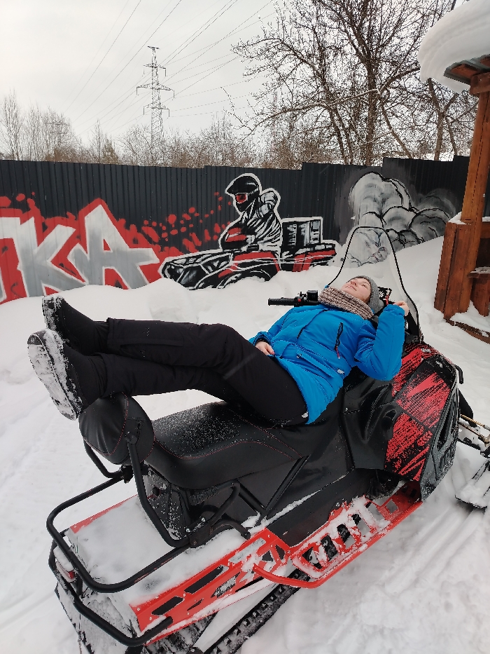 An outdoor winter scene is depicted in Chaikovsky, Russia. A single individual is positioned supine on a black and red snowmobile, which is partially covered in snow. The person is dressed in a blue winter jacket, black pants, dark boots dusted with snow, a grey knitted hat, and a patterned light-colored scarf. Their legs are elevated, with their boots resting on the snowmobile's rear passenger backrest, while their arms are bent with hands near their head. The snowmobile features red graphics on its side panel and a clear windscreen. The ground surrounding the snowmobile is covered in white snow. In the background, a dark grey corrugated metal fence is visible, adorned with red and white graffiti including stylized letters that appear to be "IKA" and a graphic depiction of a person in a helmet on a snowmobile-like vehicle. Beyond the fence, bare deciduous trees, power lines, and a tall metal electricity pylon are present under an overcast sky. A wooden structure, likely part of a shelter or play equipment, topped with a thick layer of snow, is situated on the far right.