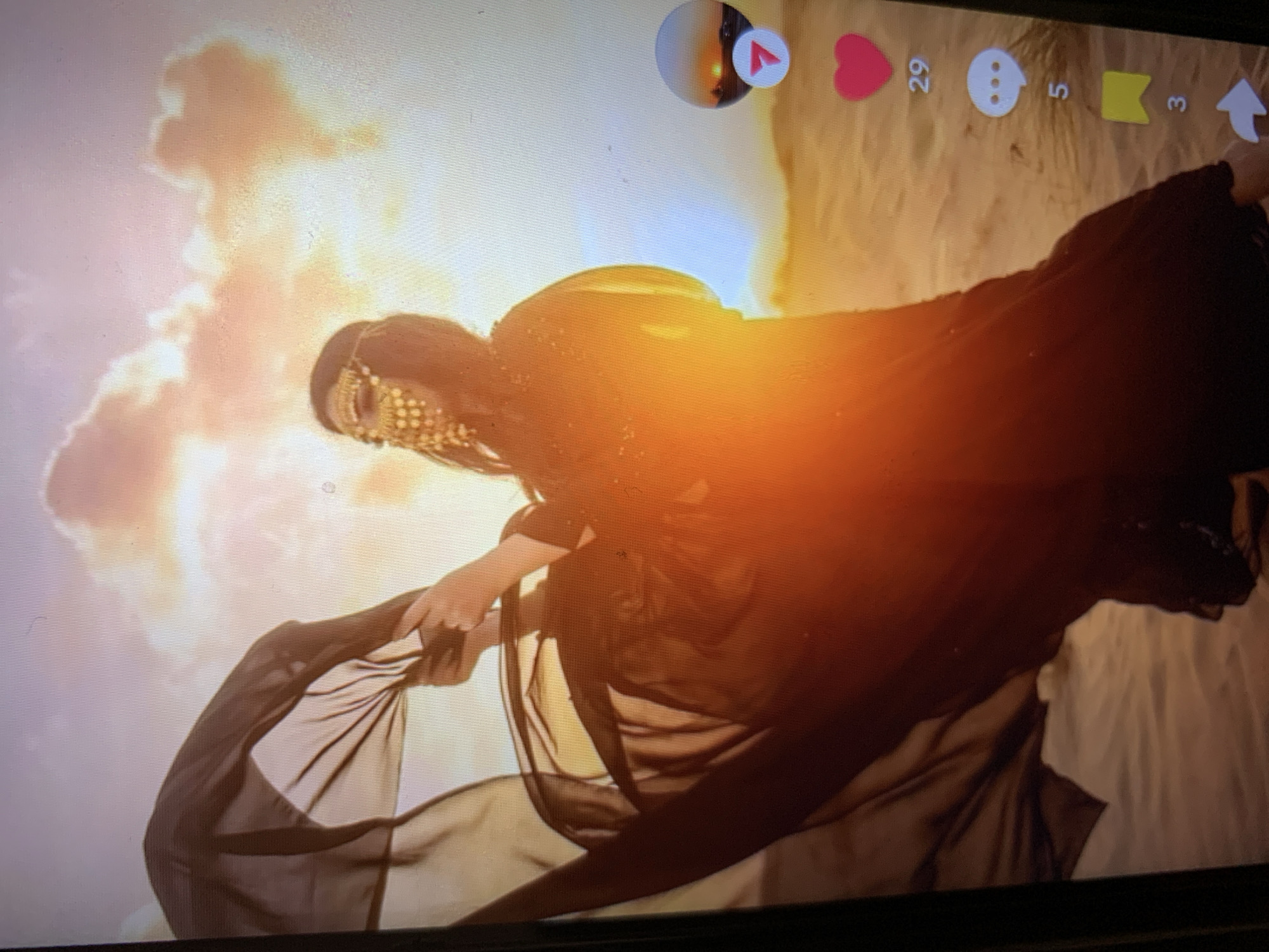 The image shows a person dressed in traditional Middle Eastern attire, including a flowing black garment and a golden headdress that partially covers their face. The person is standing in a desert landscape, with sand dunes and a dramatic sunset in the background. The sky is filled with colorful clouds, suggesting either sunrise or sunset. The person is holding a sheer black fabric, which is billowing in the wind, creating a sense of movement and drama. The image appears to be a screen capture from a social media platform, as indicated by the presence of icons for likes, comments, shares, and other interactions on the right side of the frame. The location is specified as the city of Lymanka, Ukraine.