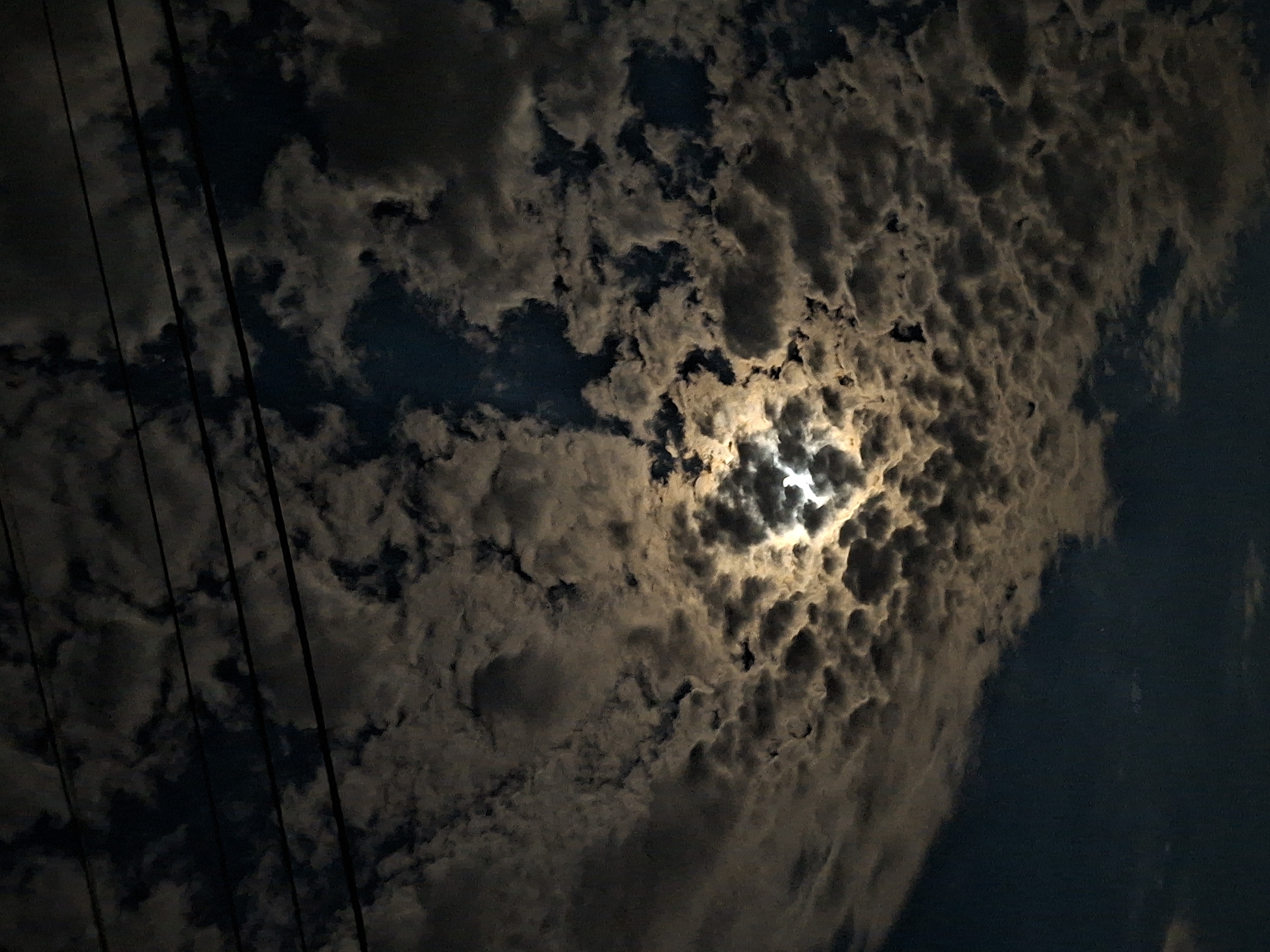 The image depicts a night sky filled with clouds in Galabovo, Bulgaria. A bright light, possibly the moon, shines through a break in the cloud cover, illuminating the surrounding clouds with a golden glow. Electrical power lines are visible in the left portion of the frame.