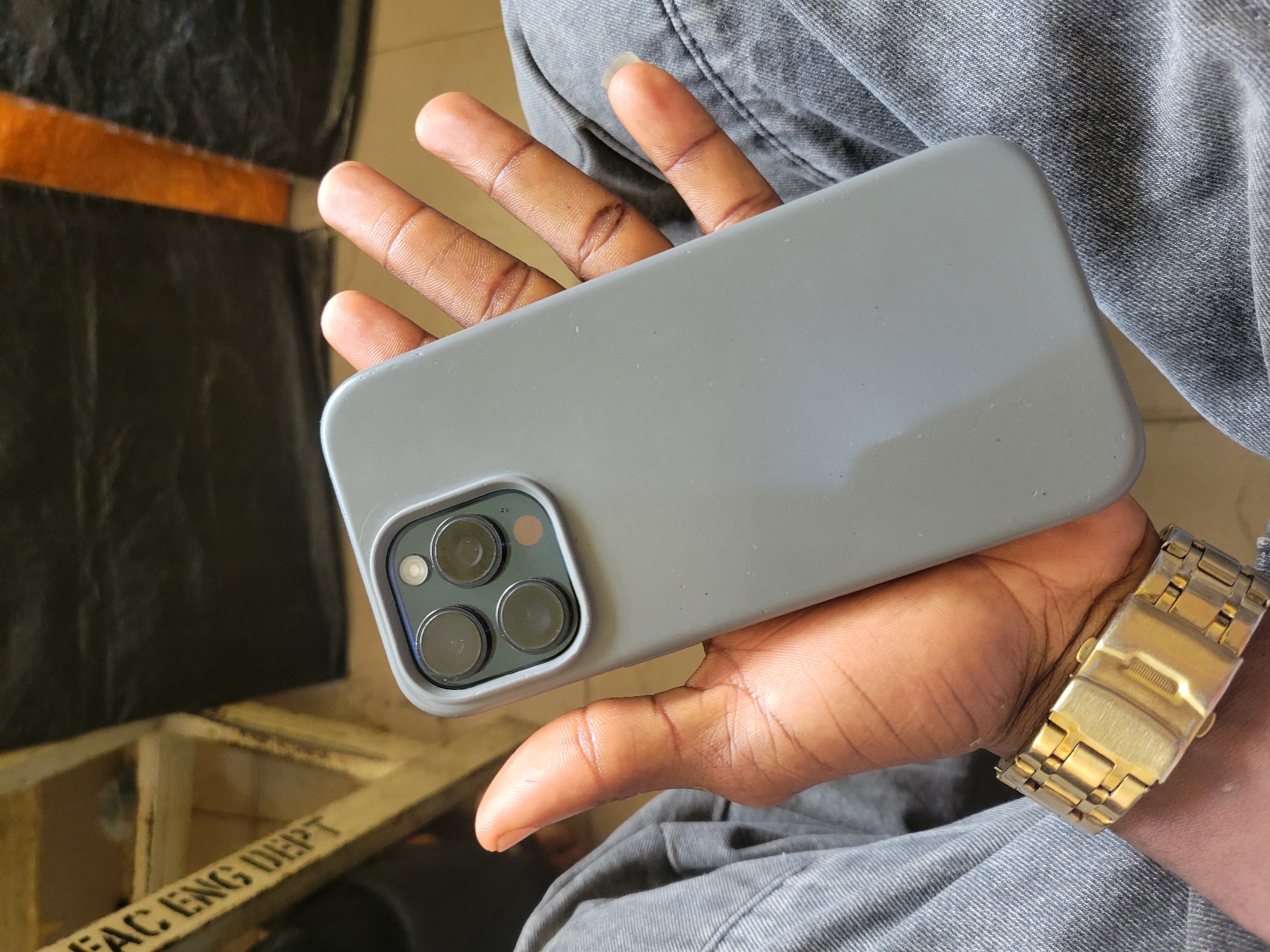The image shows a close-up, overhead view of a person's hands holding a smartphone with a gray case. The phone has a distinctive triple-lens camera system on the back. The person's skin tone suggests they are of African descent. They are wearing a gold-colored watch on their left wrist. The background is out of focus but appears to be a mix of dark, possibly tarp-like material, and a wooden structure with some text visible. The overall scene is casual, possibly taken indoors or in a shaded outdoor area, given the lighting. The text on the wooden structure appears to say "FAC ENG DEPT". The location context is Ukukwa, Nigeria.