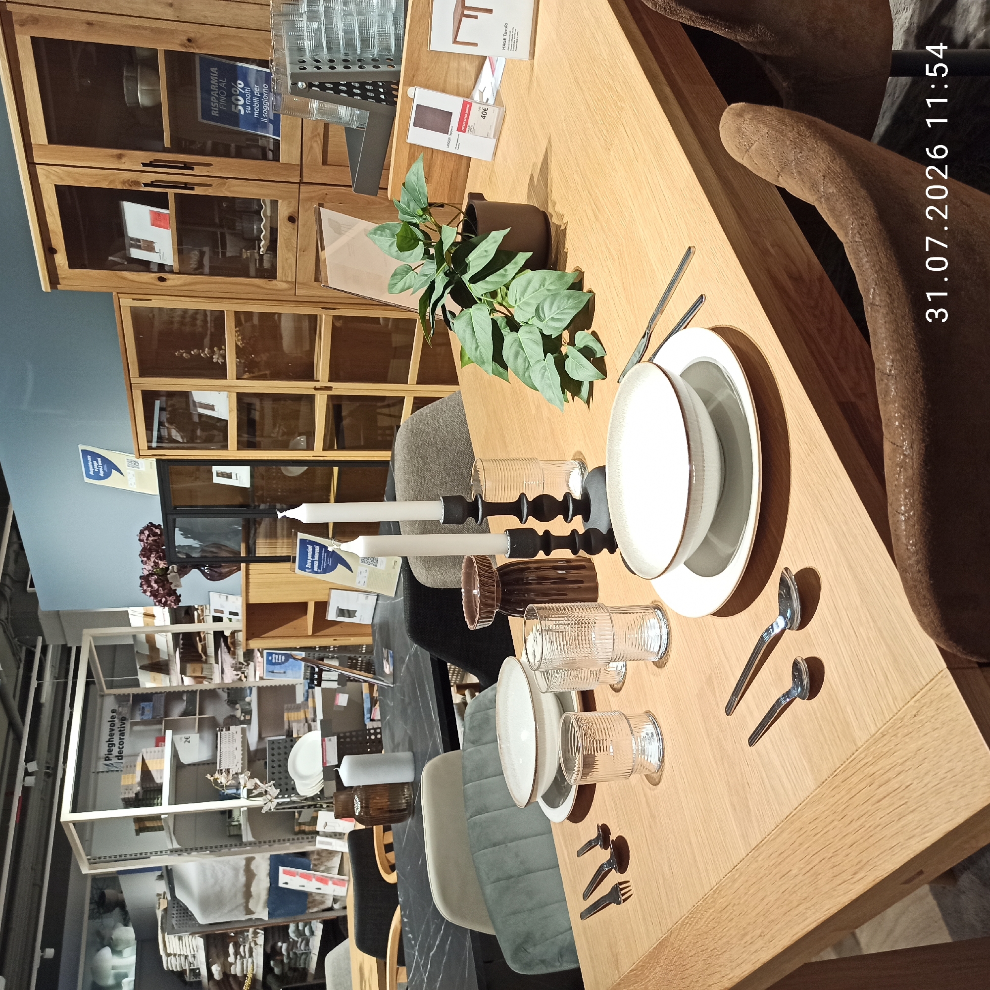 This is a photograph of a dining table set in what appears to be a showroom or retail environment. The table is made of light-colored wood and is set for at least two people with place settings that include a bowl, plate, fork, and spoon. There are also two ribbed glasses and a knife on the table. A dark brown vase with a textured pattern and two white taper candles in a black candlestick holder are positioned near the center of the table. A potted plant with large green leaves sits to the right of the table, near a stack of glasses in a metal holder. In the background, there are display shelves filled with various items, including boxes and decorative objects. A tall, wooden cabinet with glass doors is visible to the right, showcasing more items. Two chairs are partially visible at the table, one with a light-colored upholstered back and another with a brown, textured fabric. The overall lighting suggests an indoor setting. A timestamp overlay at the bottom right of the image reads "31.07.2026 11:54". The image is taken from a slightly elevated angle, looking down at the table.