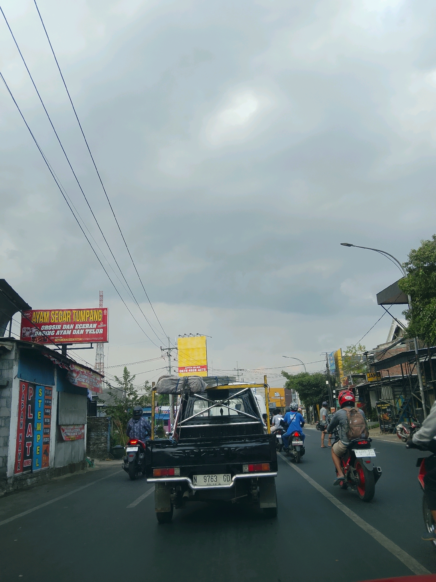 The image depicts a street scene in Madyopuro, Indonesia, on a cloudy day. The foreground is dominated by the rear of a black pickup truck, its license plate reading "N 9763 GD". To the left, a building with a blue facade displays signs, including one that reads "AYAM SEGAR TUM...". Further down the street, several motorcycles and their riders are visible. One motorcyclist in a blue shirt and helmet is moving away from the camera, while another rider with a backpack and red helmet is facing away. The street is wet, suggesting recent rain. Overhead, power lines stretch across the frame. The environment is characterized by various buildings, street lights, and trees lining the roadside, indicating an urban or semi-urban setting.