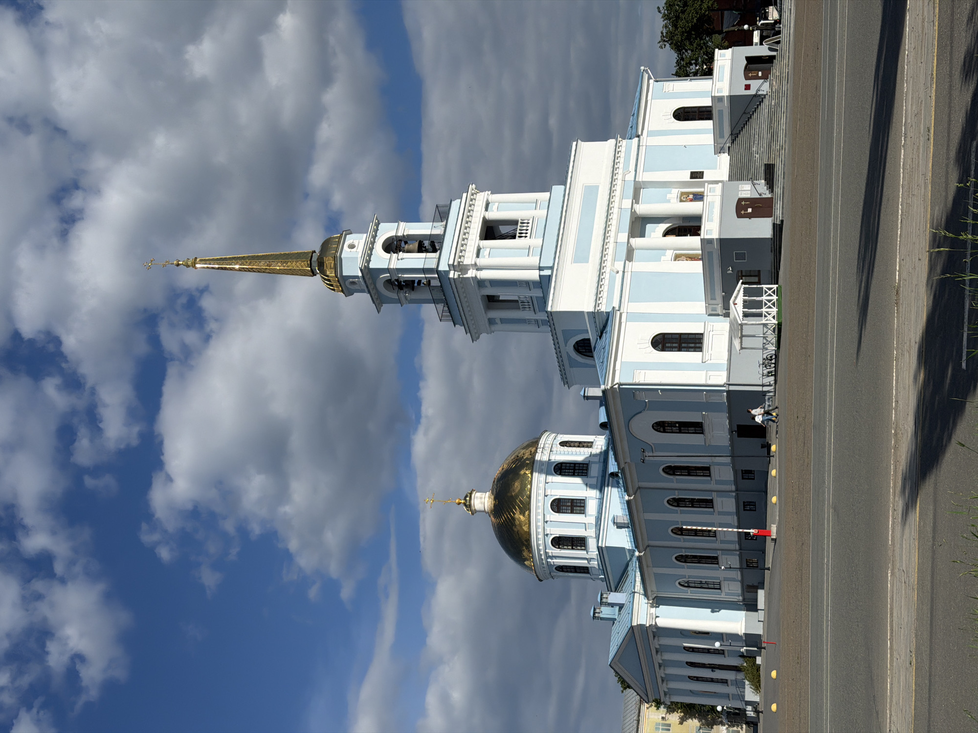 The image displays the Blagoveshchensky Cathedral (Annunciation Cathedral) in Votkinsk, Udmurtia, Russia. The large, classical-style church is predominantly light blue and white, featuring a prominent golden dome topped with an Orthodox cross on the left and a tall, multi-tiered bell tower with a pointed golden spire, also crowned with an Orthodox cross, on the right. The bell tower contains visible bells within its upper open section. The building's facades include numerous arched windows with dark frames and white pilasters, along with sections featuring classical columns. A grand stone staircase ascends to the main elevated entrance, where two religious icons are affixed to the wall above the steps. Additional ground-level entrances are visible. A small, white, open-sided gazebo-like structure stands near the base of the main stairs on the left.
The sky above is partially cloudy, with patches of clear blue interspersed with scattered white and grey cumulus clouds. In the foreground, an asphalt road runs horizontally across the bottom, separated by a light-colored paved area from the building. Yellow spherical bollards are spaced along the edge of the paved area, and a red-and-white striped barrier post is centrally located. Long, dark shadows are cast across the foreground pavement, suggesting direct sunlight from a high, slightly right-of-center source relative to the viewer. Two individuals are walking on the paved area near the right side of the building's entrance, and one indistinct person is visible further left near the building. Sparse green grass is visible at the very bottom edge of the frame.