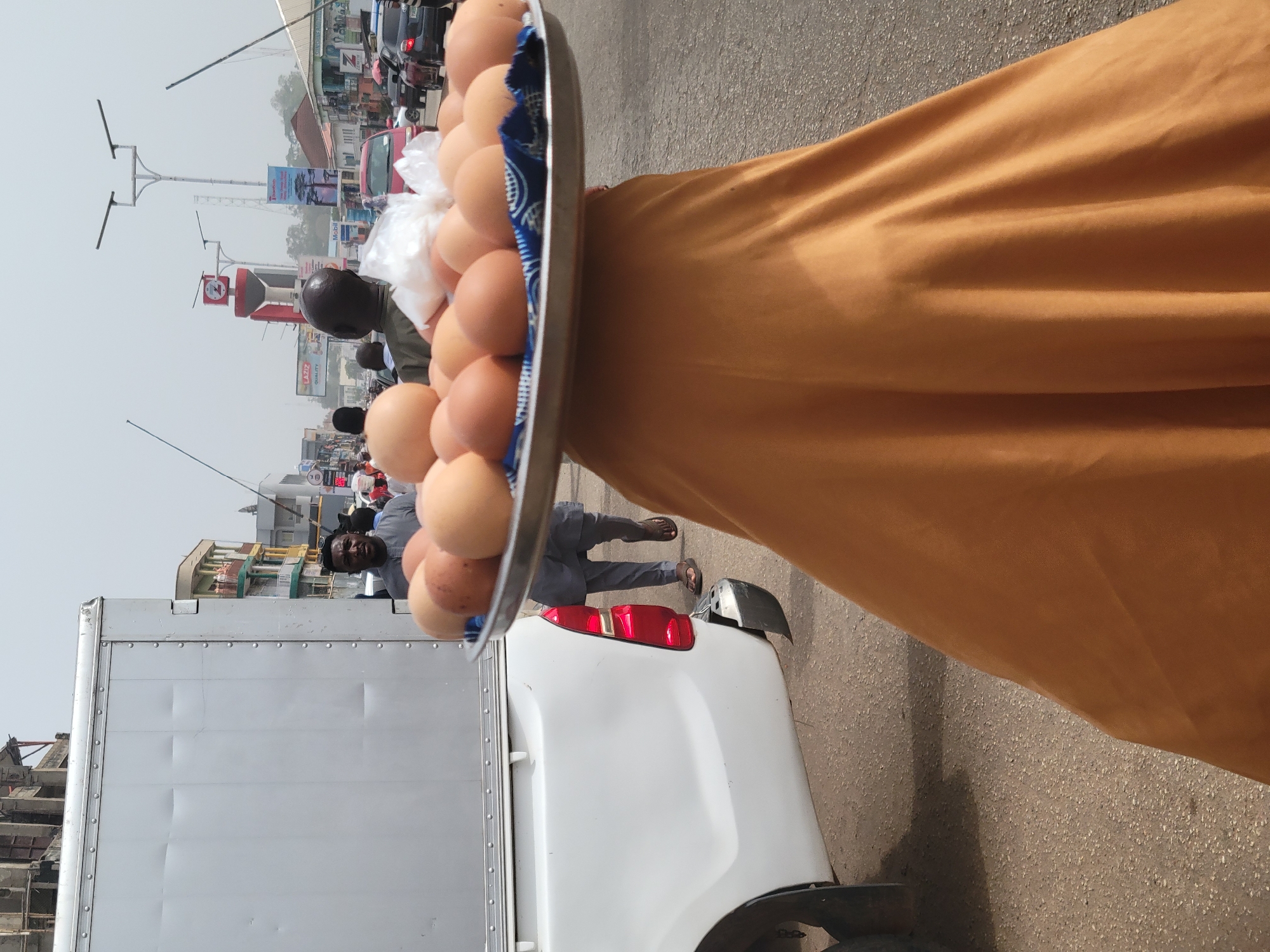 This street-level image captures a vibrant, sunny day in Jos, Nigeria, from the first-person perspective of a street vendor holding a large metal tray filled with numerous light brown eggs. The tray is partially lined with a blue and white patterned fabric and also holds a small white plastic bag. The vendor's mustard yellow garment is visible along the right side of the frame.
In the mid-ground, a white pickup truck with a utility body is prominent, partially obscuring the street. Behind the truck, several people are visible: one man with dark skin and a short haircut is looking towards the camera, while another man in a grey traditional outfit is walking or standing further back, wearing sandals.
The background reveals a bustling urban street scene. Buildings of varying heights line the road, suggesting a commercial area. Traffic is present, with other vehicles visible in the distance. The street is equipped with modern streetlights featuring horizontal crossbars. Multiple billboards and signs are visible, including one for "MOBIL" (a gas station), another reading "LA BIZ QUALITY," and a red structure with a visible clock or number "2." The bright, clear sky indicates daytime. The scene depicts everyday life and commerce in a Nigerian city.